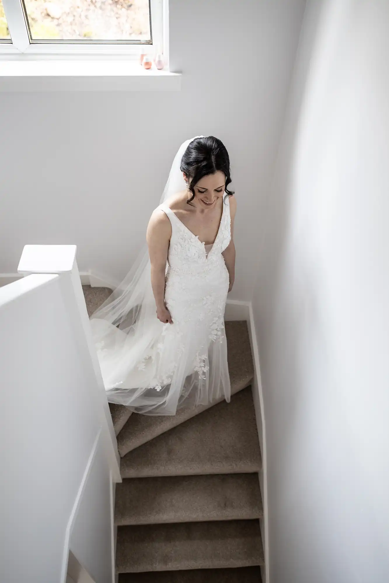 A bride in a white gown and veil descends a carpeted staircase in a well-lit room.