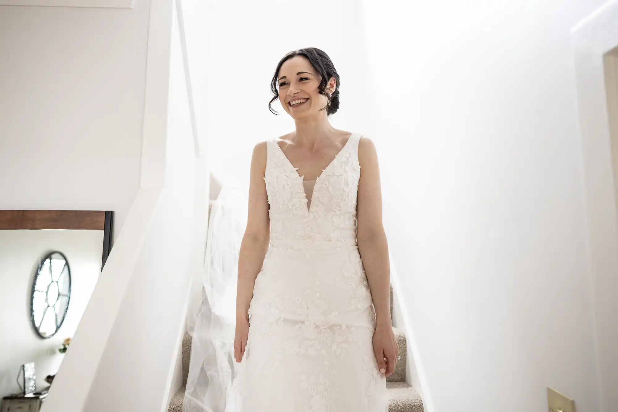 A woman in a white wedding dress with floral details stands on a staircase, smiling.