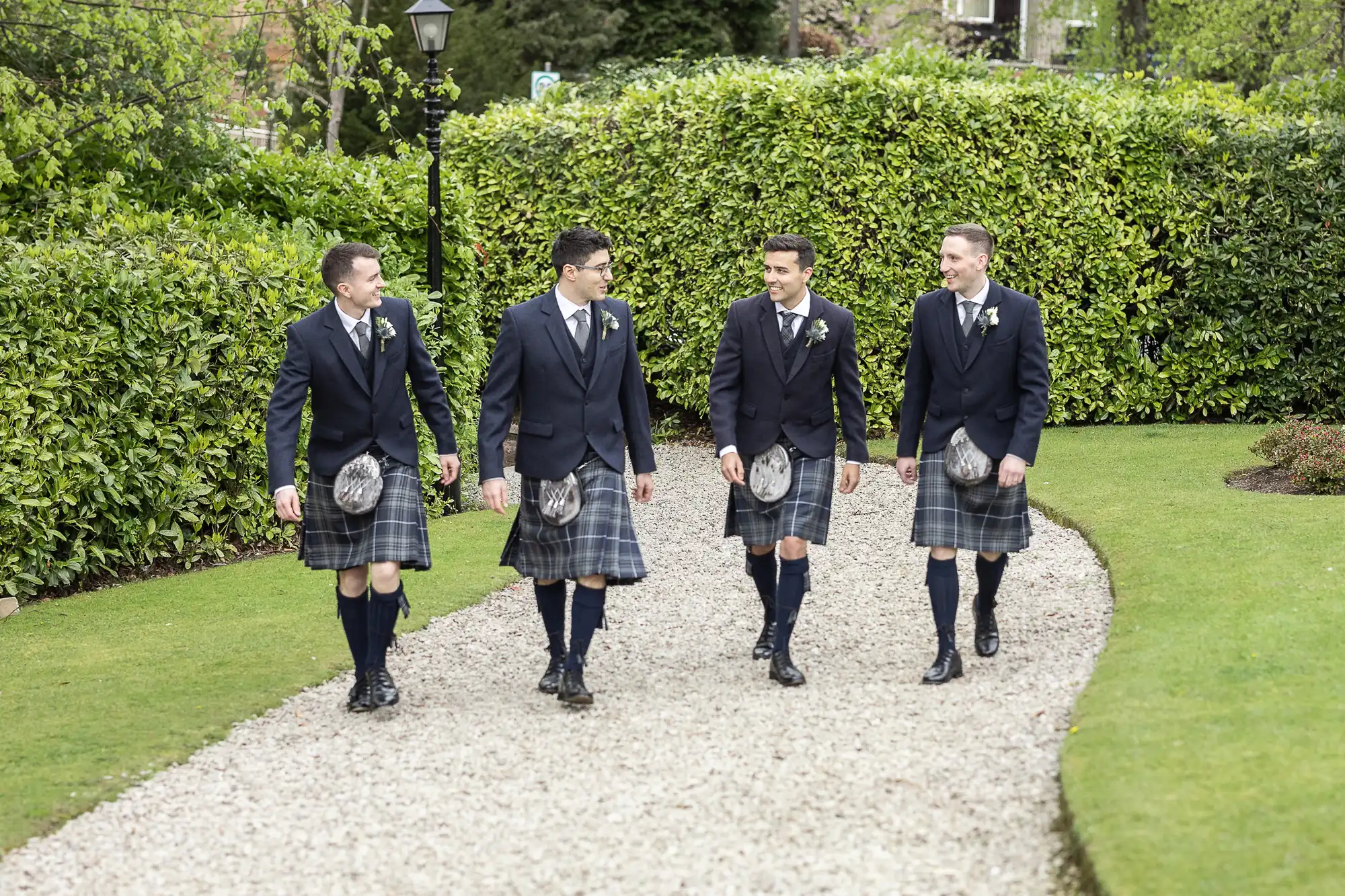 Four men in kilts and jackets walk along a gravel path lined with greenery, engaged in conversation.