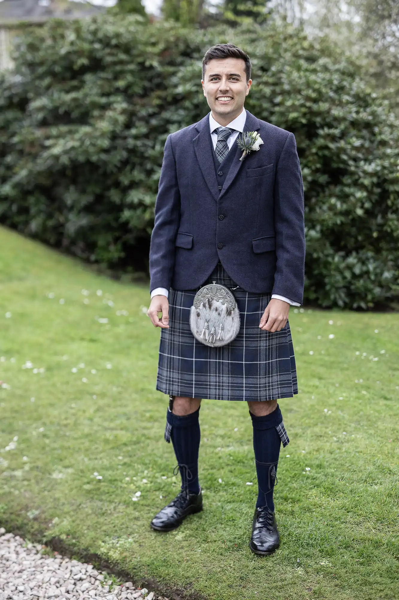 Man in a navy jacket and plaid kilt stands on grass, wearing a sporran, long socks, and a tie. Bushes are in the background.