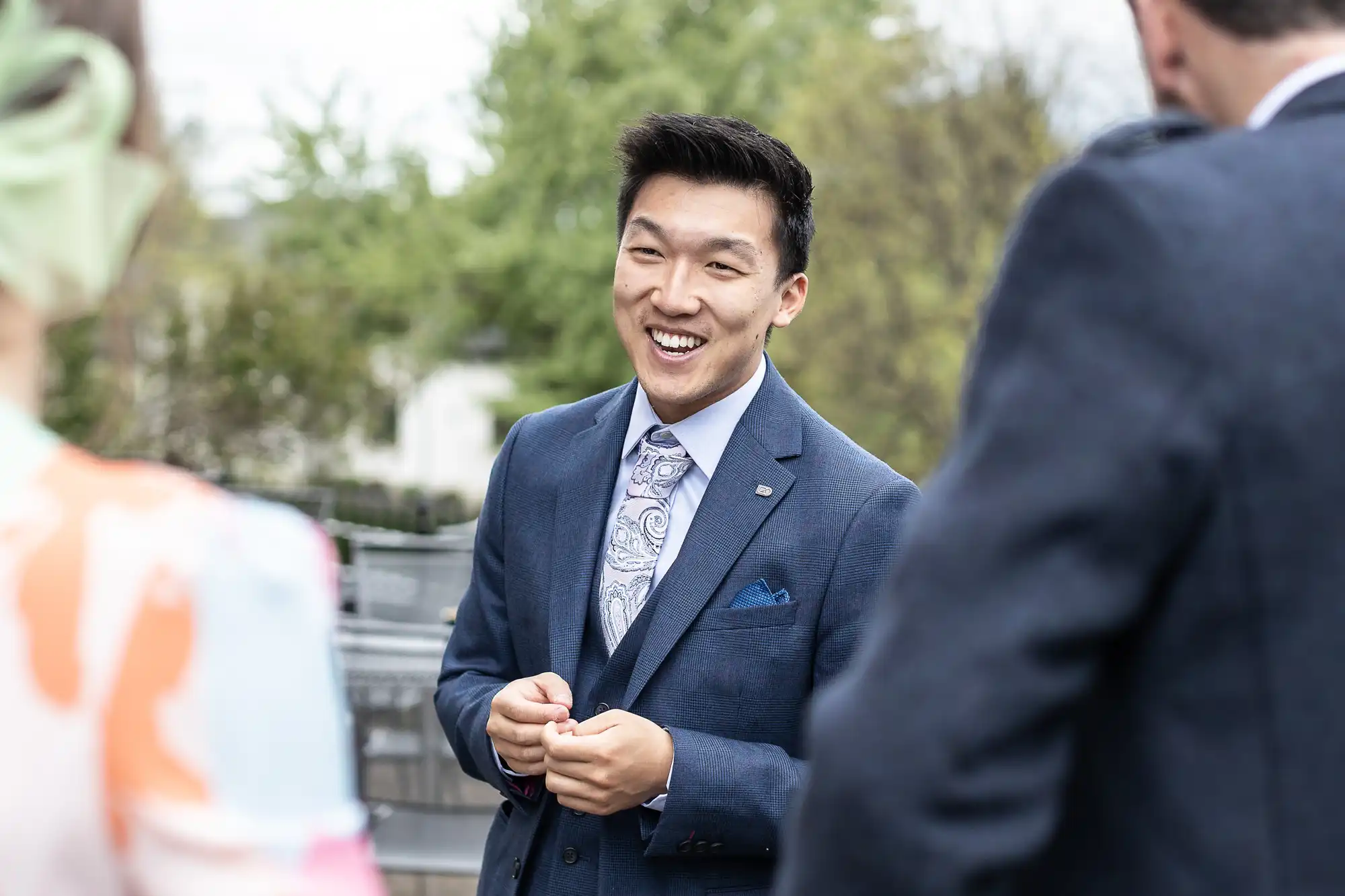 A man in a suit smiles and talks with two people outdoors, with trees in the background.