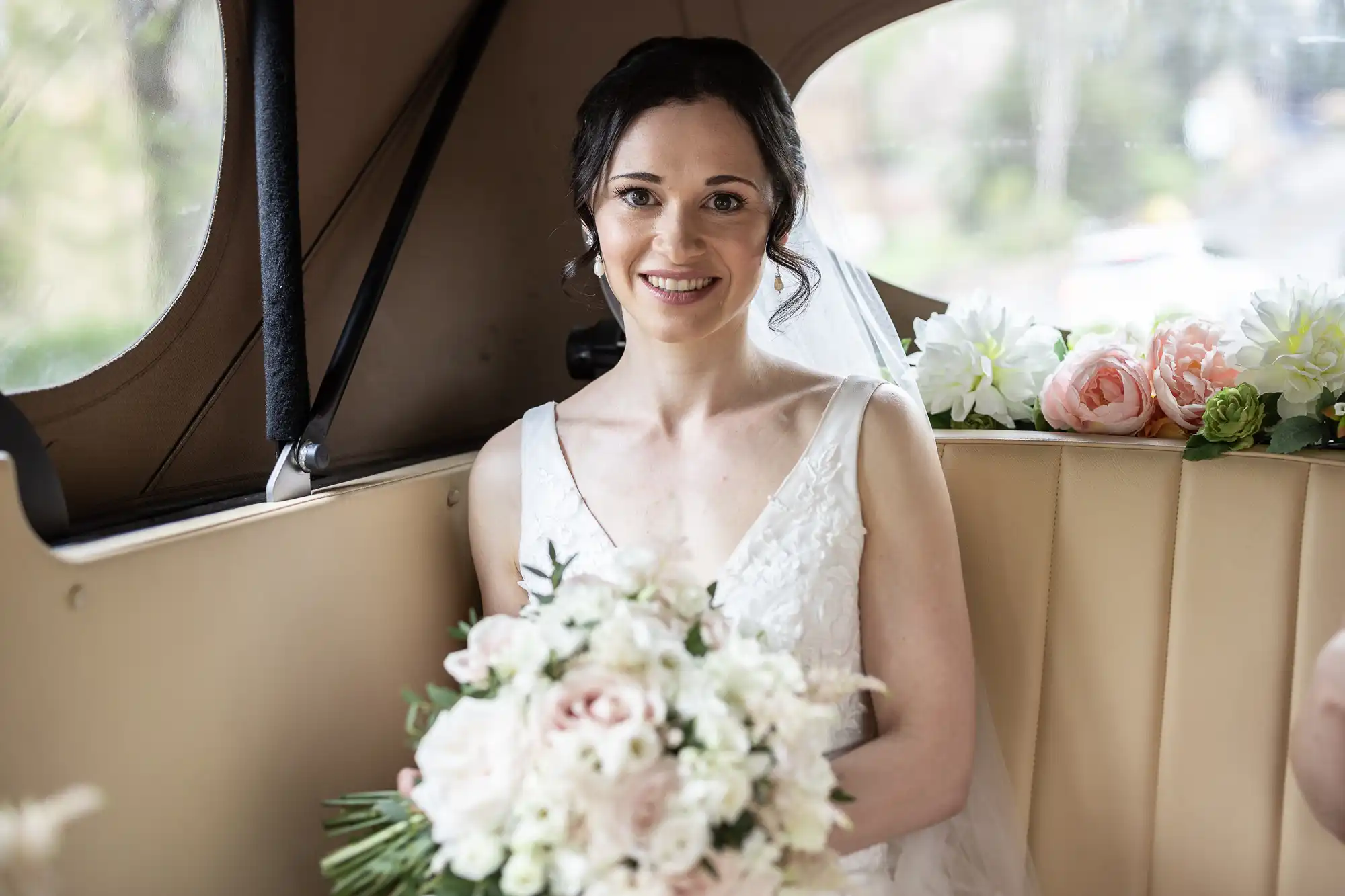 Bride in a white dress holding a bouquet of white and pink flowers, sitting in the back of a car decorated with floral arrangements.