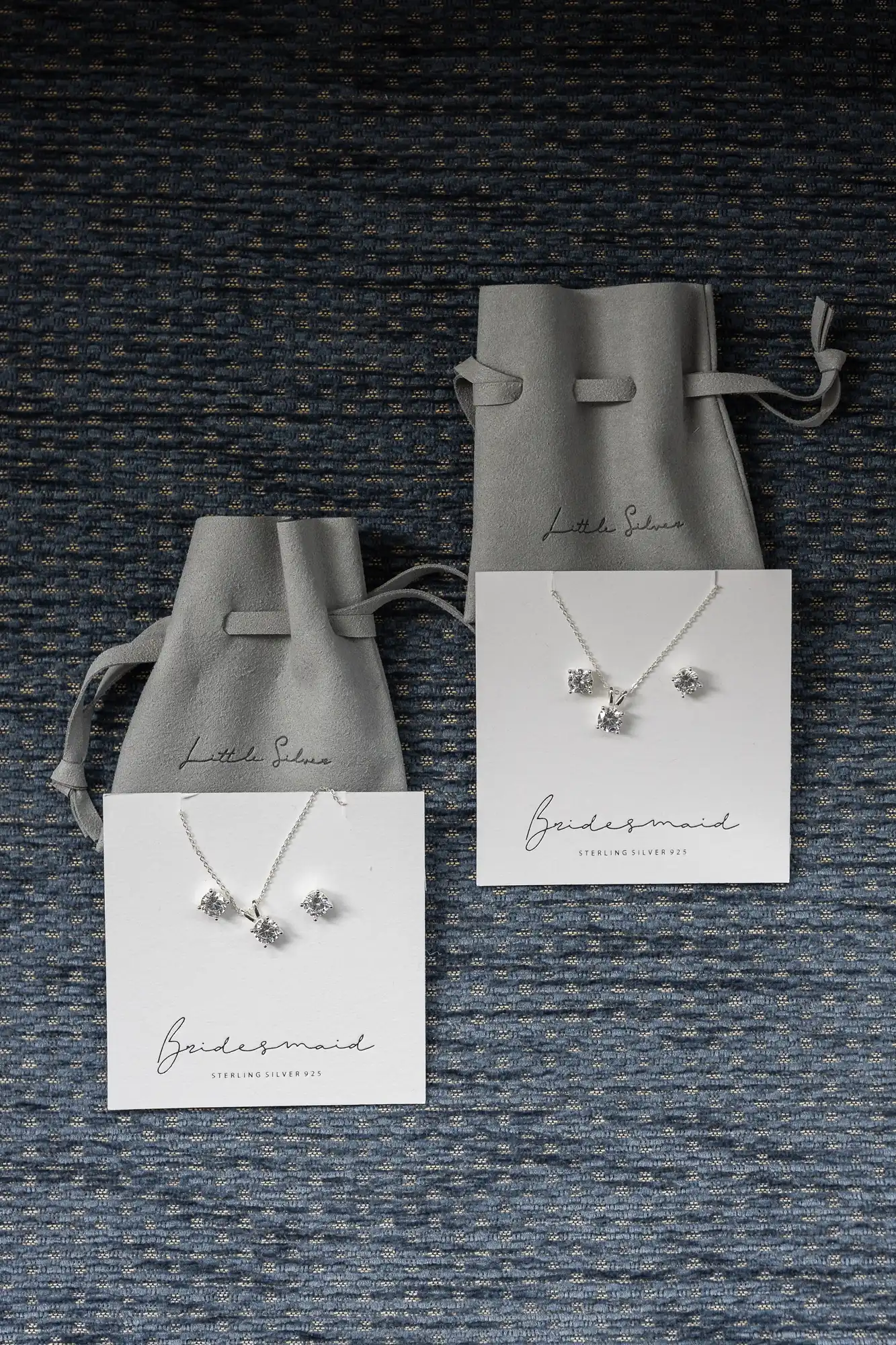 Two jewelry sets labeled "Bridesmaid" with silver necklace and earrings on white cards, placed on a textured dark blue surface, accompanied by gray pouches labeled "Little Silver.