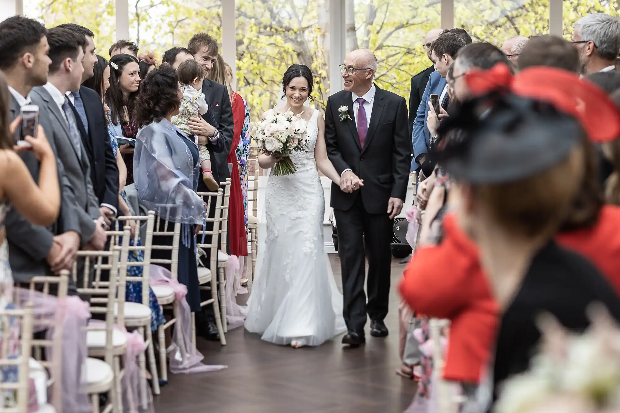 A bride in a white dress walks down the aisle with an older man in a suit. Guests seated on both sides watch and take photos in a venue with large windows and trees visible outside.