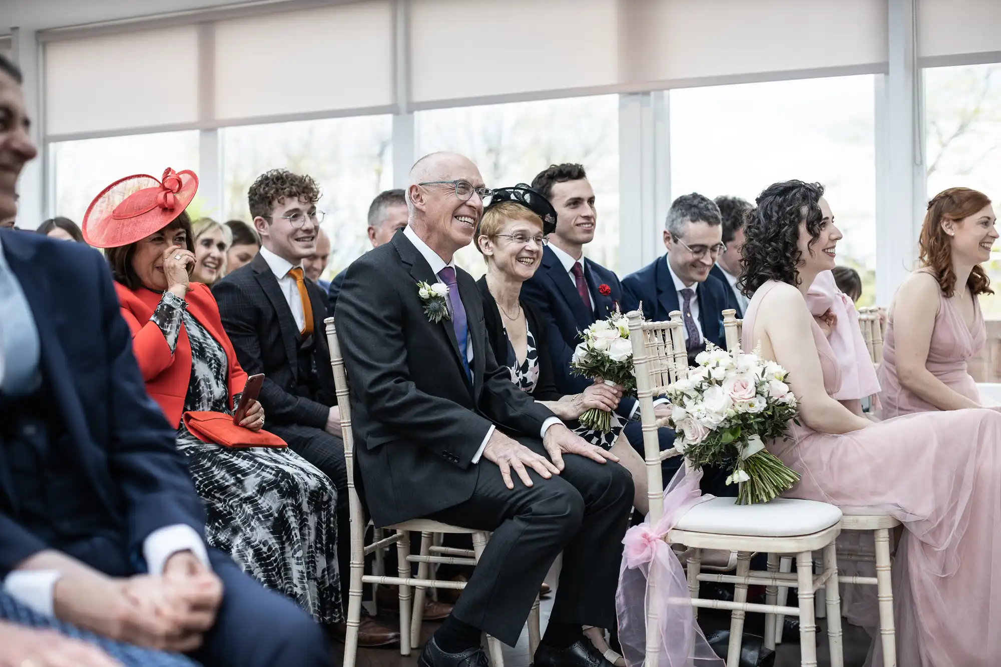 Guests sitting and smiling at a wedding ceremony, some holding bouquets.
