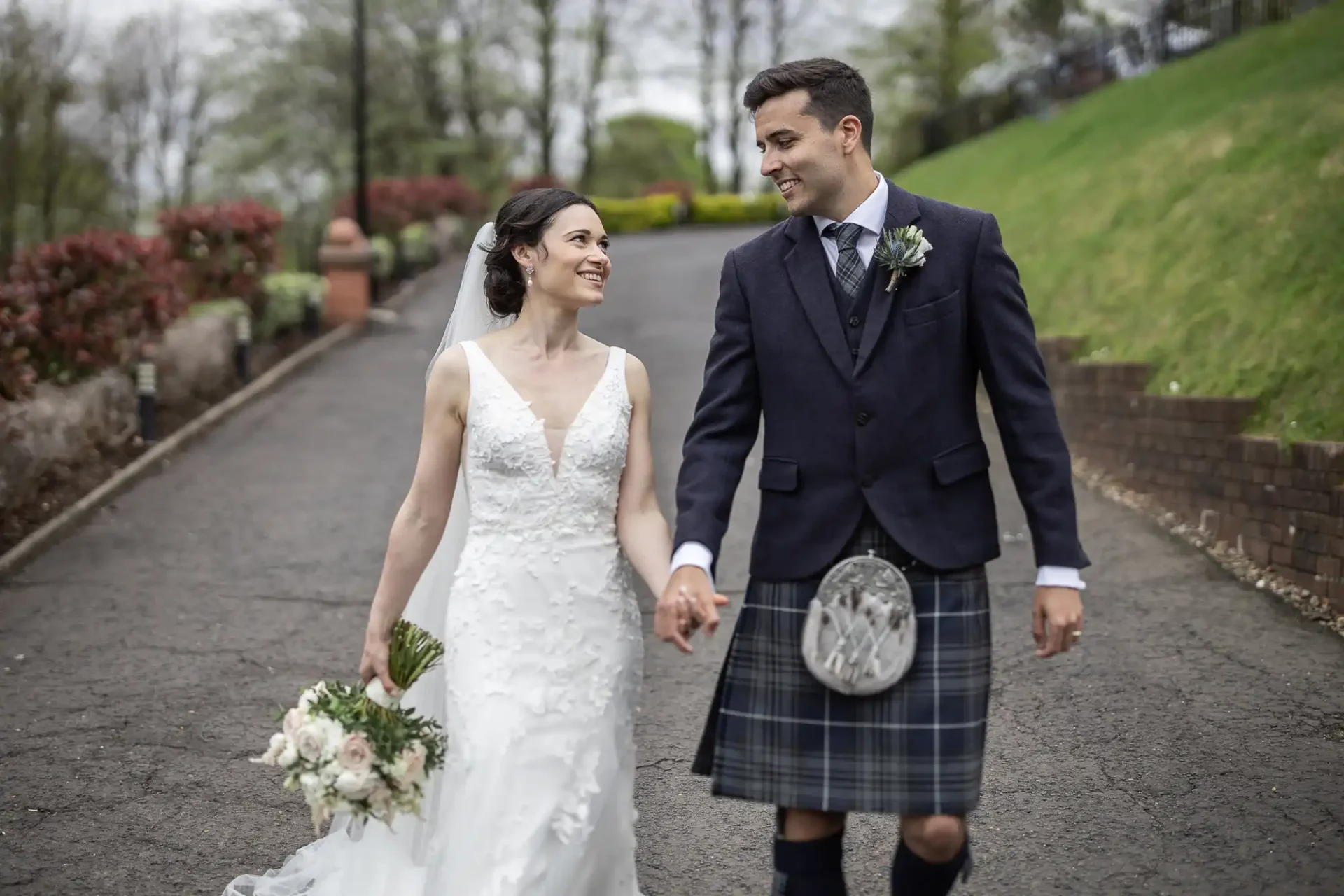 Sherbrooke Castle Hotel wedding photographer image of a bride in white dress and groom in a kilt walking on a tree-lined path, holding hands and smiling at each other.