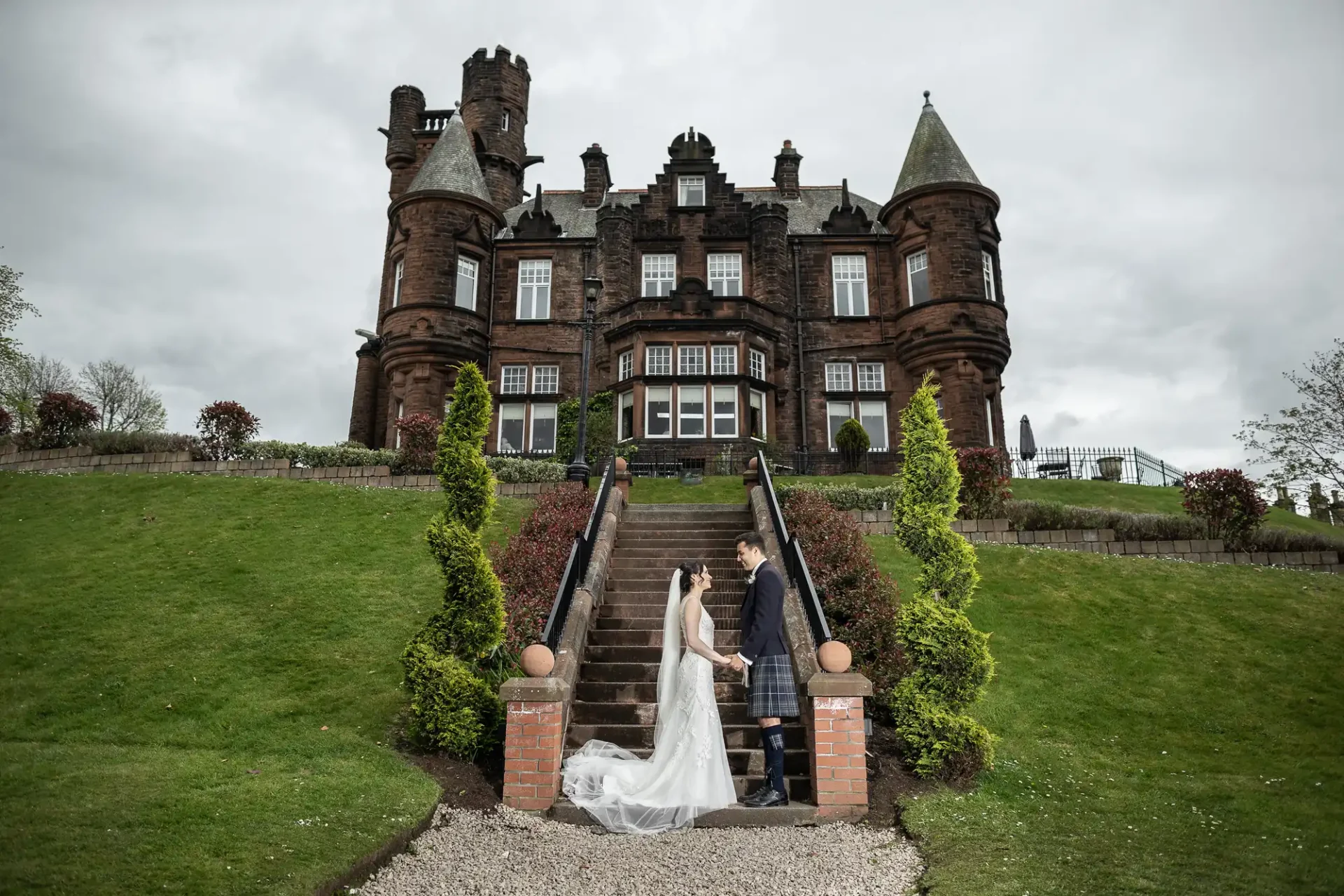 A bride and groom stand on steps in front of Sherbrooke Castle with cones and turrets, surrounded by greenery and a cloudy sky.