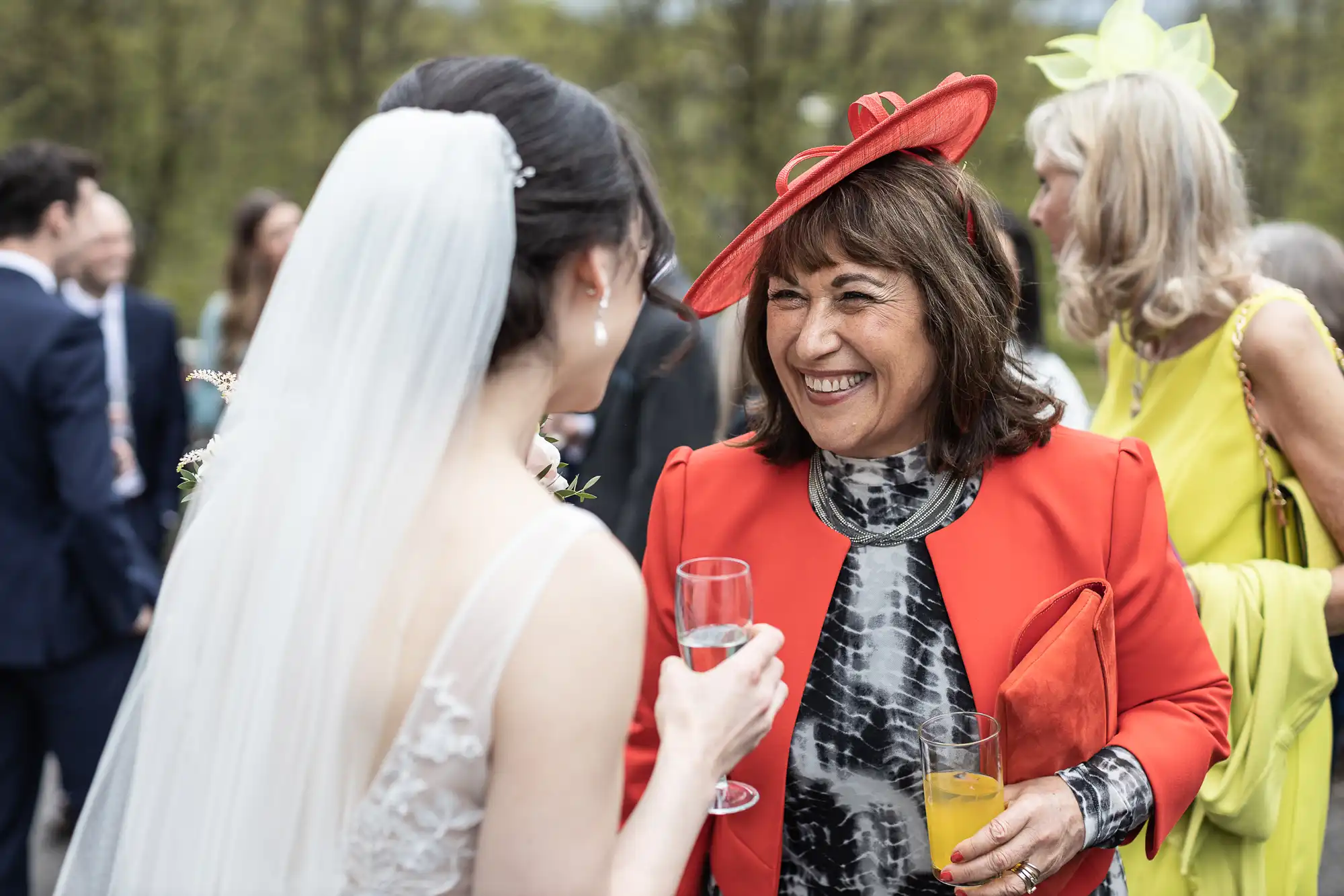 A bride in a white dress and veil talks with a smiling woman in a red outfit holding a drink at an outdoor event.