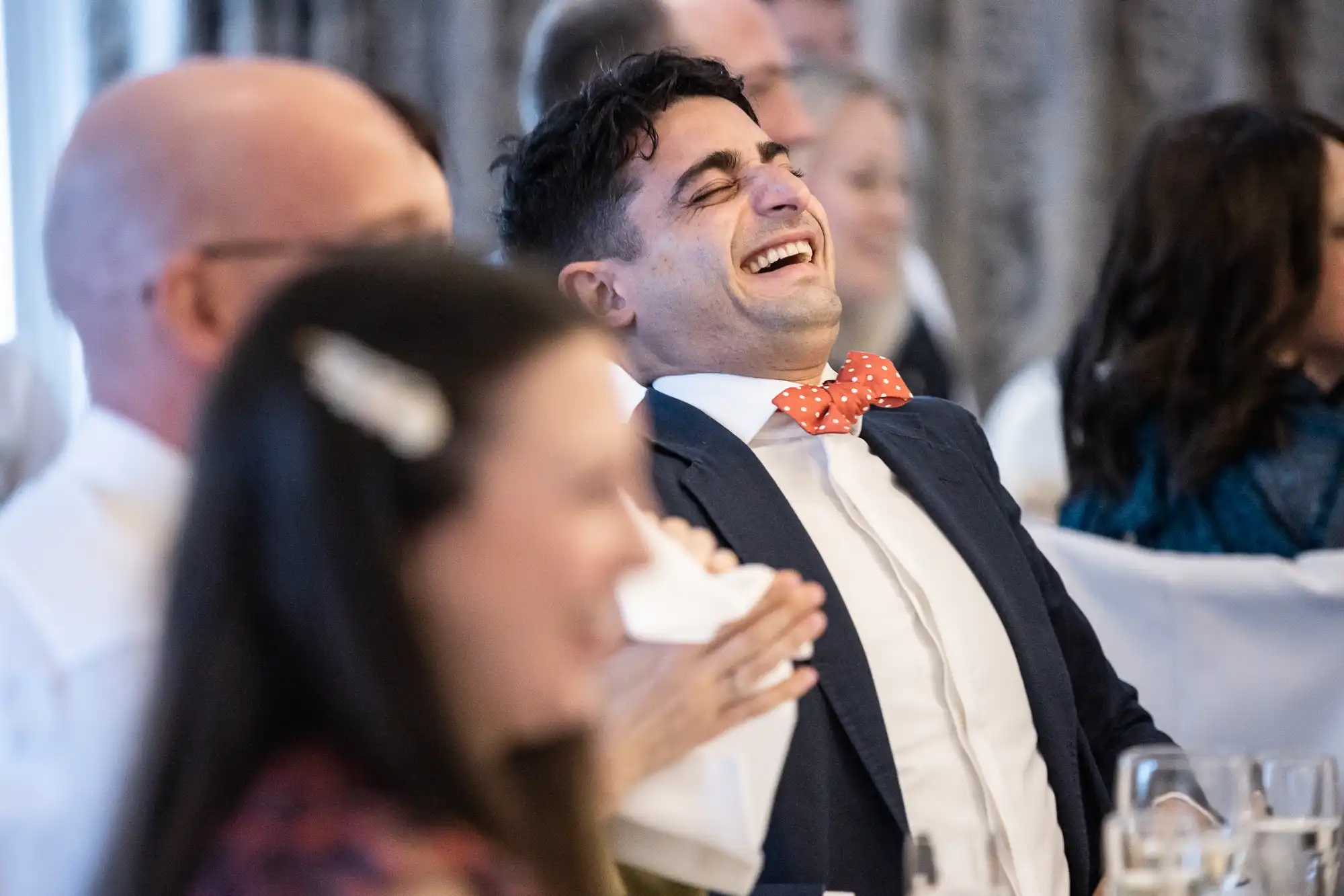Person in a suit and polka dot bow tie laughing heartily among a group of seated people at an indoor event.