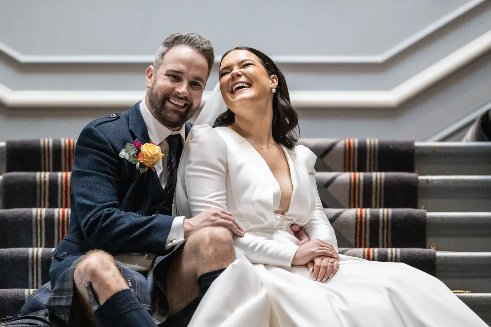Signet Library wedding photographer portrait of Helen and Daniel in Edinburgh. A joyful couple in wedding attire sitting on a staircase, the man in a kilt and the woman in a white dress, both laughing.