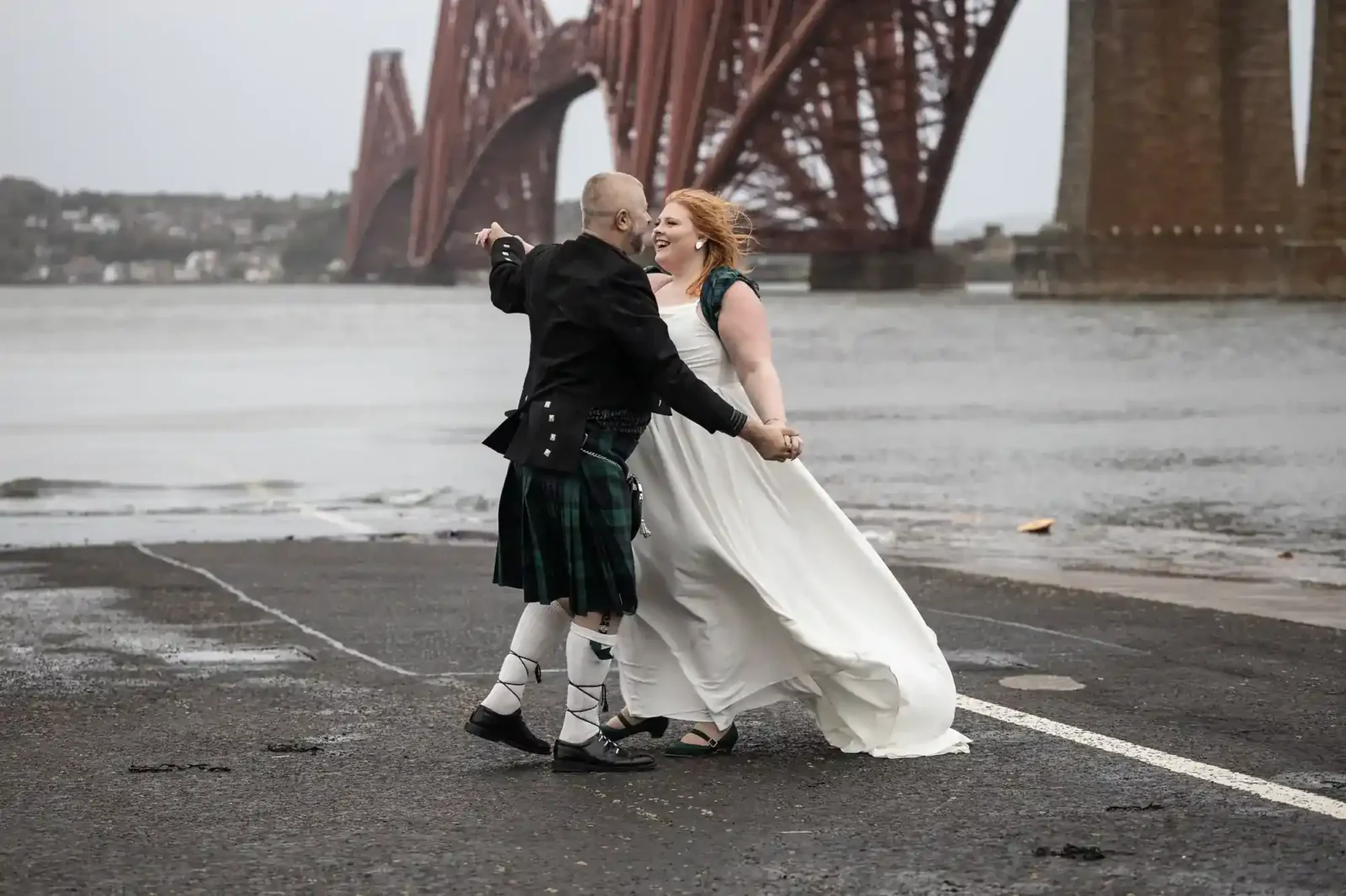 A couple in wedding attire dances on a wet pavement near a large red steel bridge, the Forth Bridge over a body of water on a cloudy day.
