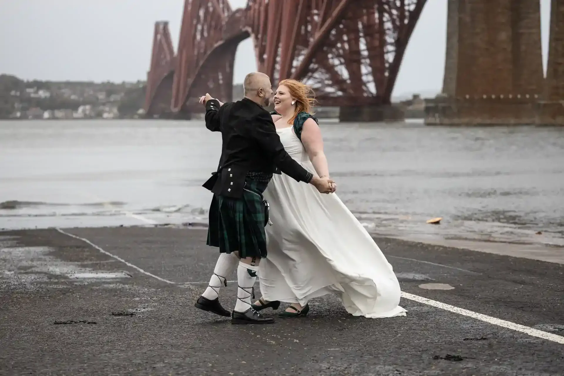 A couple in wedding attire dances on a wet pavement near a large red steel bridge over a body of water on a cloudy day.