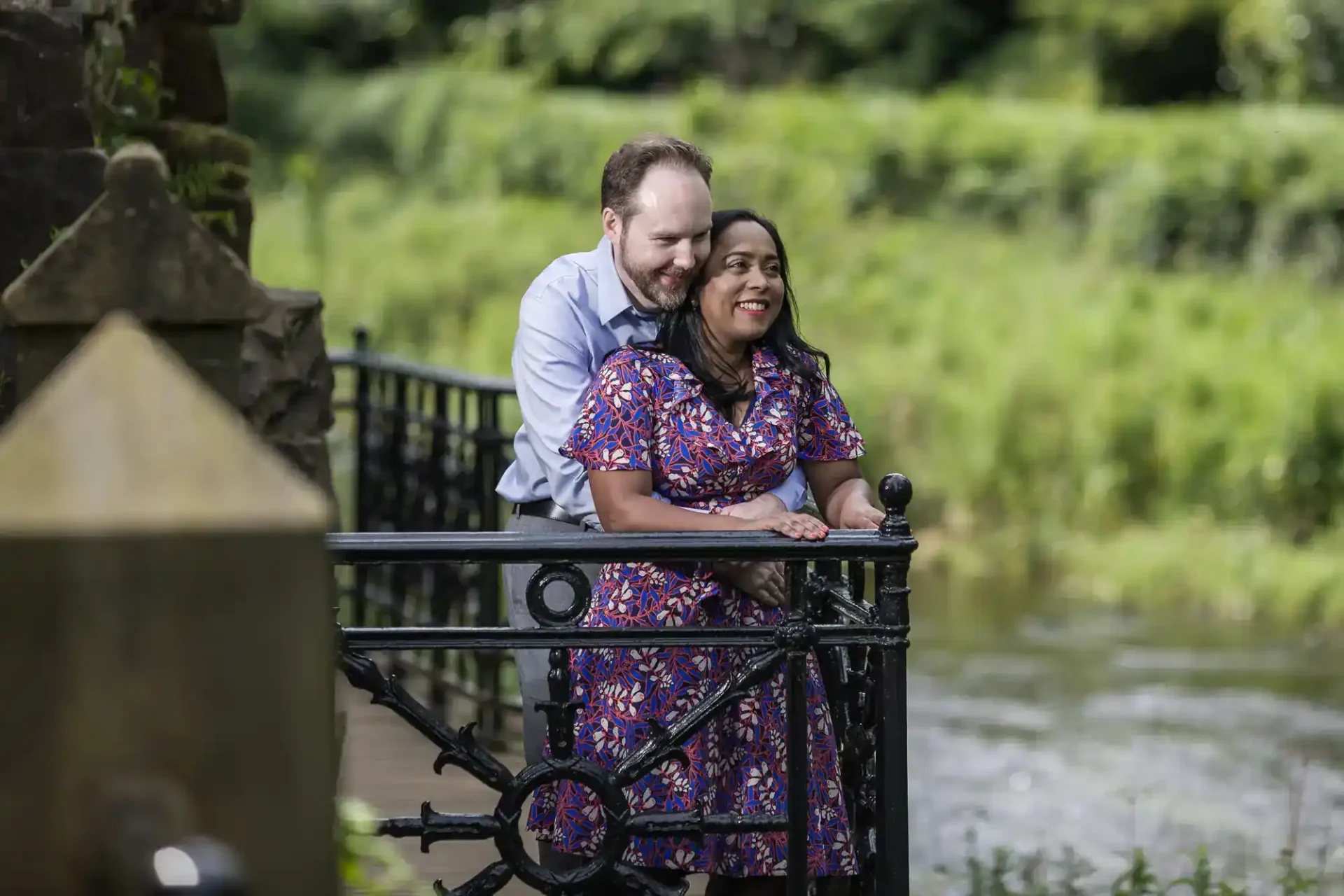 A man and woman stand together by a black metal railing overlooking a river, with greenery in the background. The man is embracing the woman from behind and both are smiling.