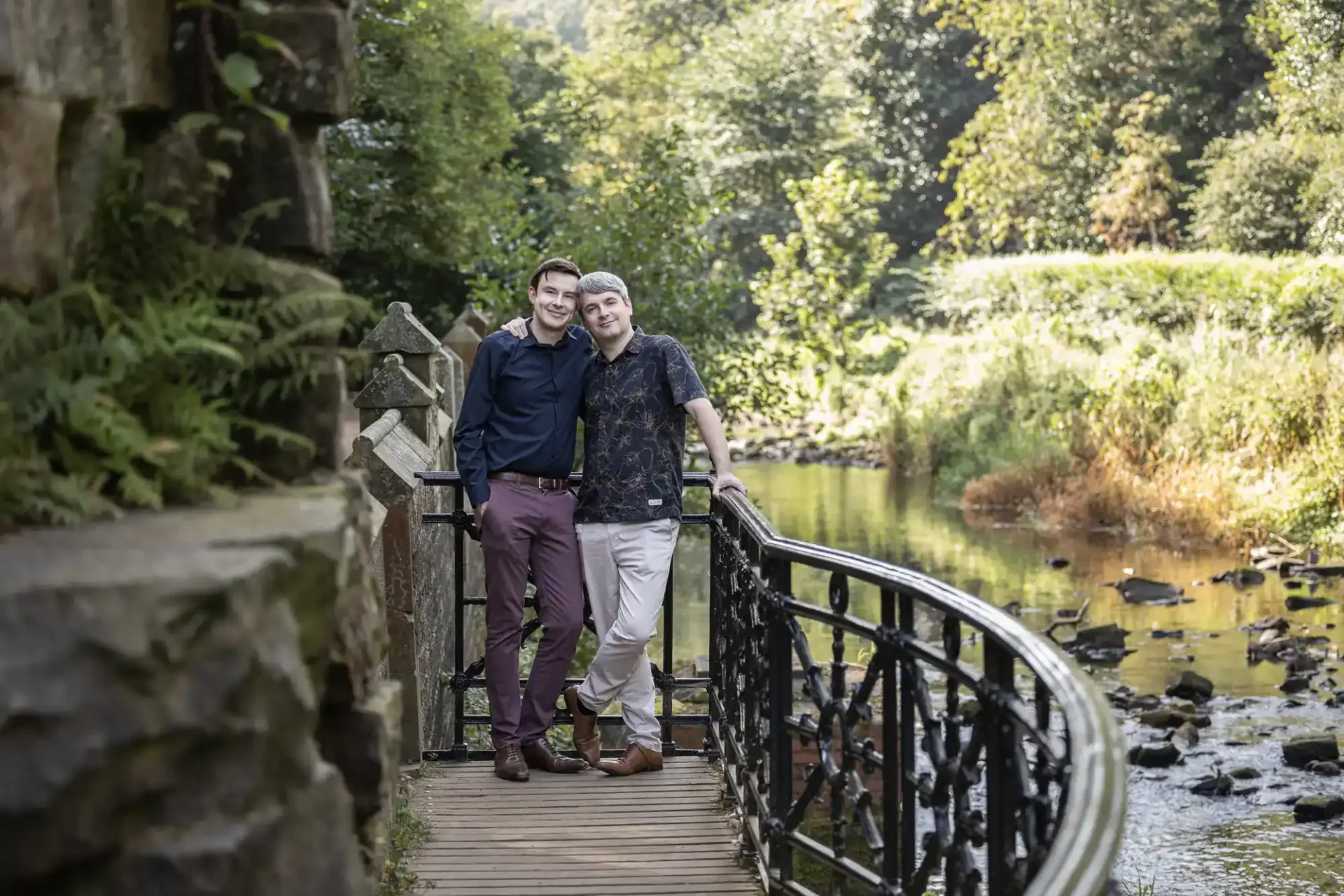 Two people stand closely together, smiling on a small bridge over a stream surrounded by greenery and trees in a park-like setting.