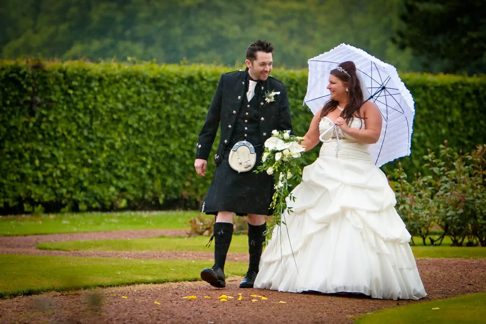 A bride in a white dress and a groom in a kilt walking under an umbrella on a rainy day, smiling as they walk on a path sprinkled with yellow petals.
