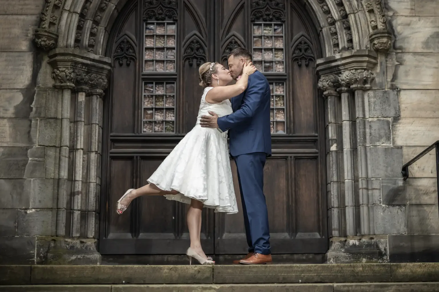 A couple, dressed in wedding attire, embraces on the steps of a gothic-style building. The bride wears a knee-length white dress, and the groom is in a navy suit.