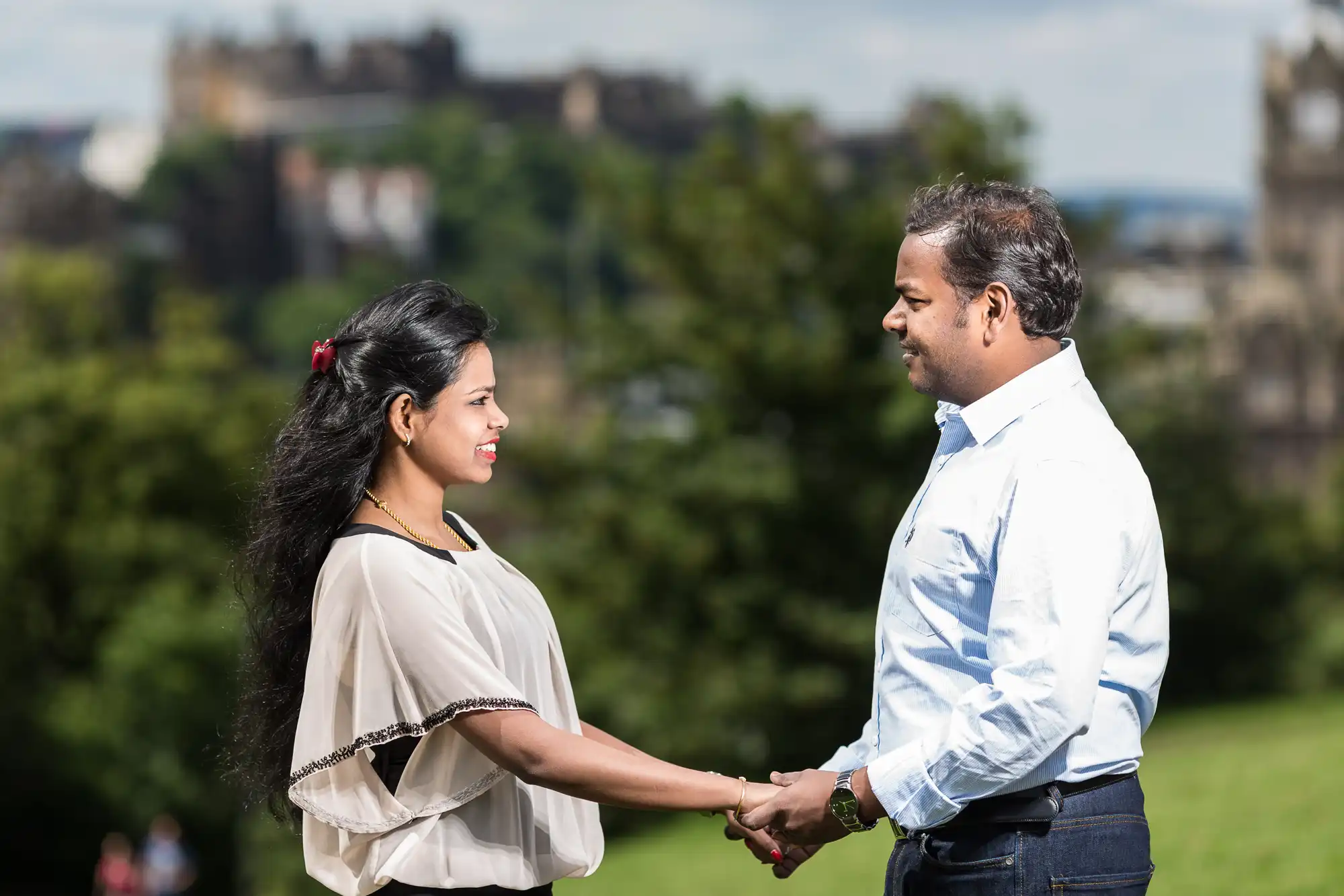 A man and a woman stand outdoors, holding hands and looking at each other. Blurred greenery and buildings are visible in the background.