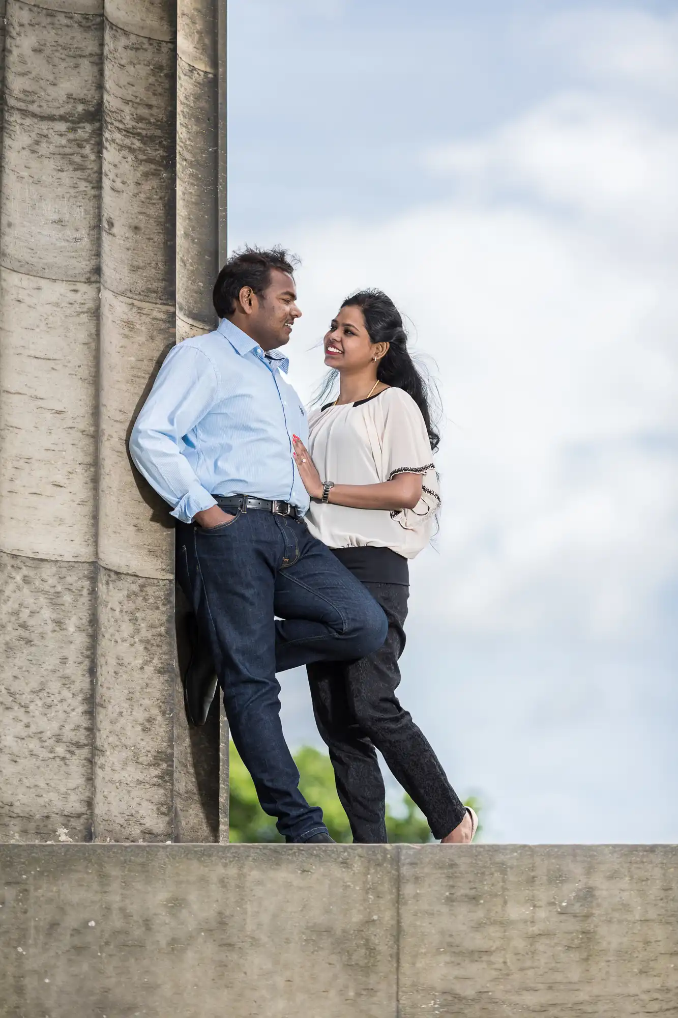 A man and a woman stand next to a pillar, casually chatting and smiling at each other, with a partly cloudy sky in the background.