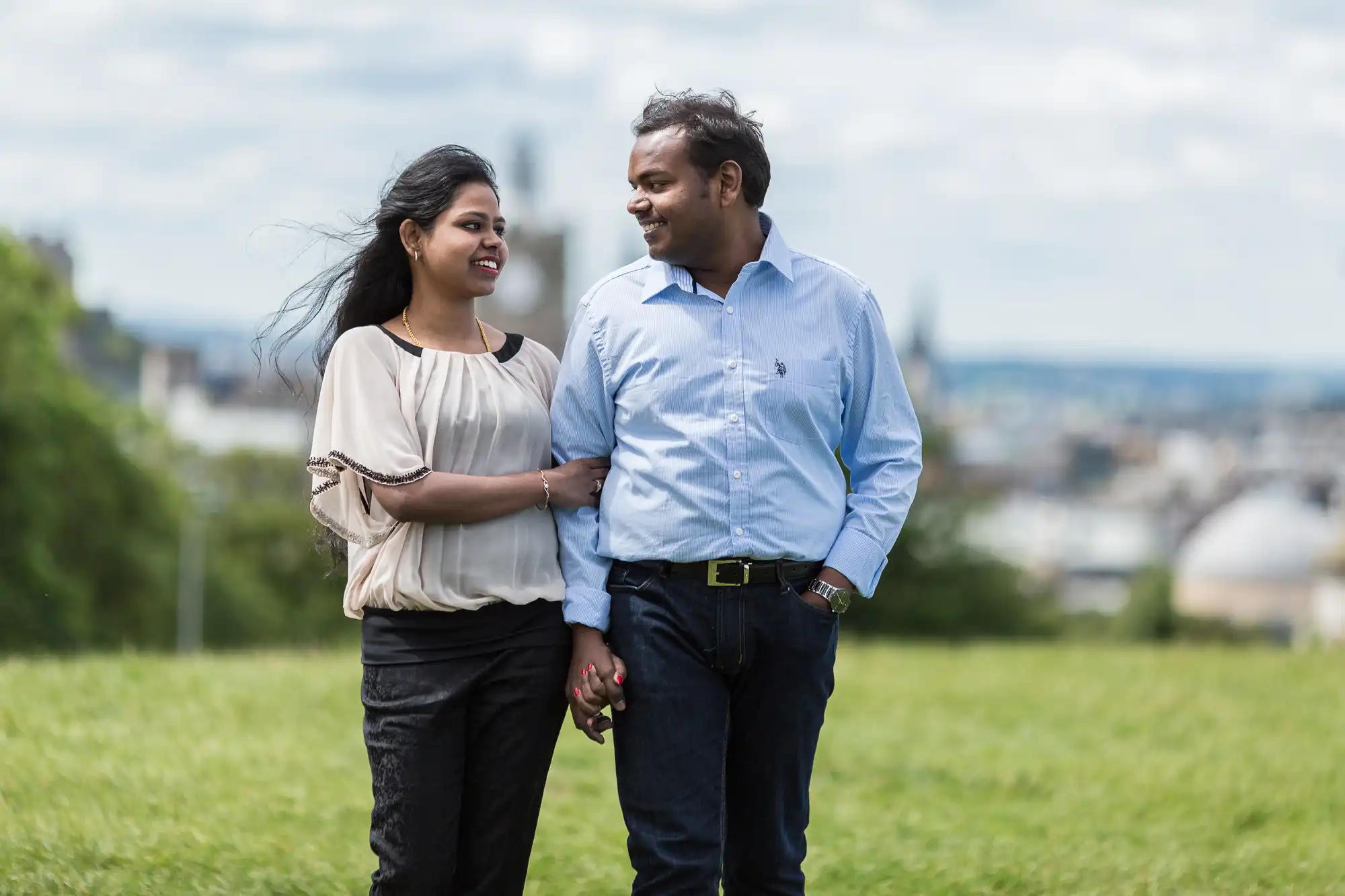 A couple walks hand in hand through a grassy field, smiling at each other with a blurred cityscape in the background.