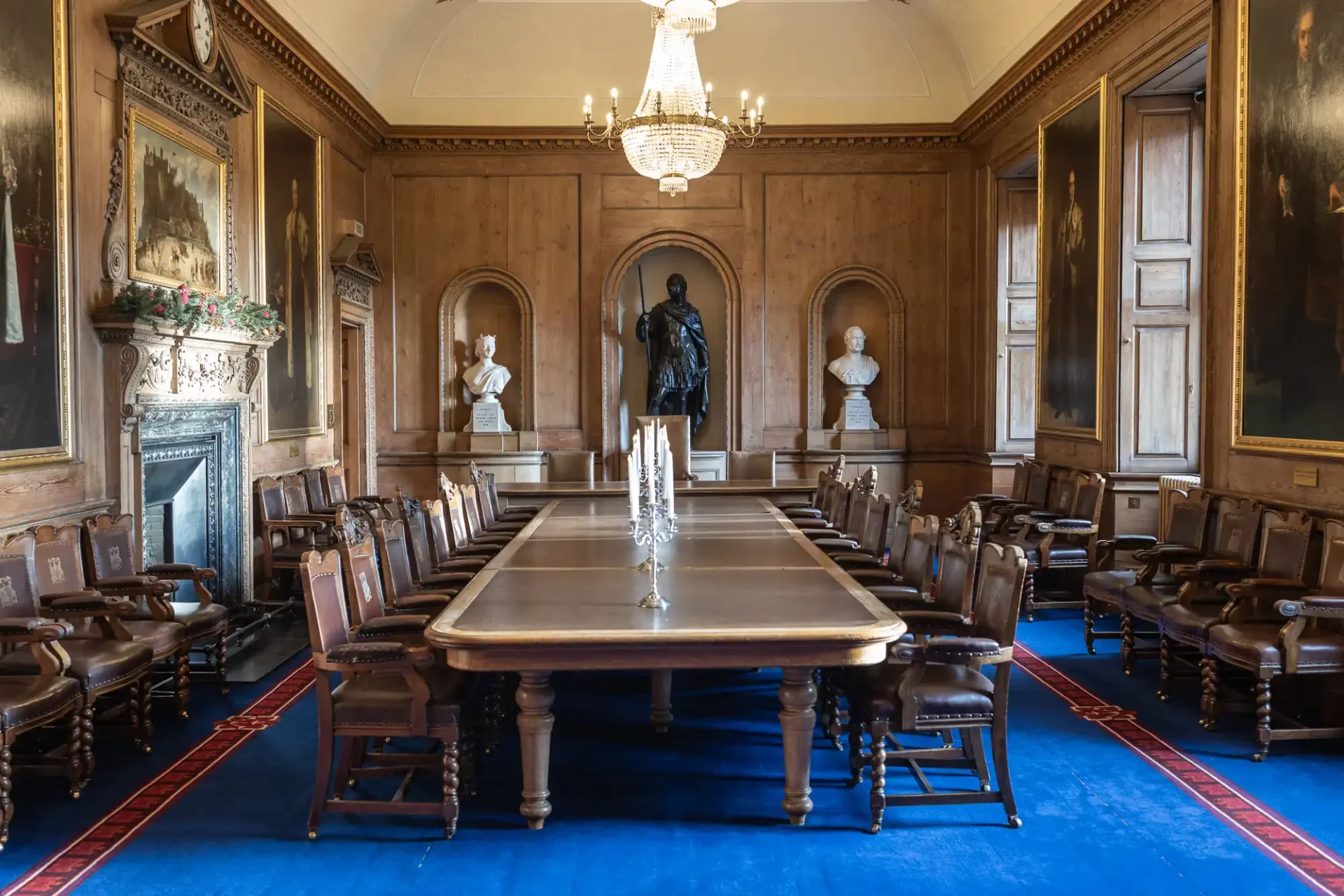 Diamond Jubilee Room at Edinburgh City Chambers. A formal dining room with a long wooden table, surrounded by chairs. Portraits adorn the wood-paneled walls, and a chandelier hangs from the ceiling. Busts are displayed in the background.