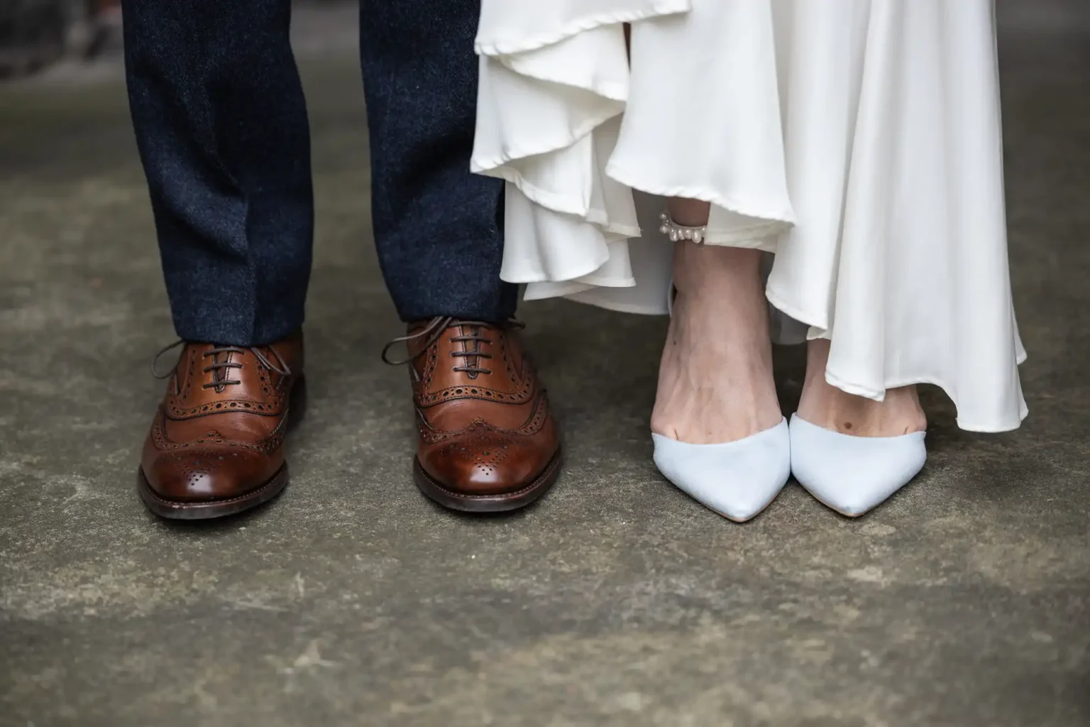 Close-up of a bride and groom's feet. The groom wears brown leather brogues, and the bride wears white pointed heels. Her white dress drapes slightly above the ground.