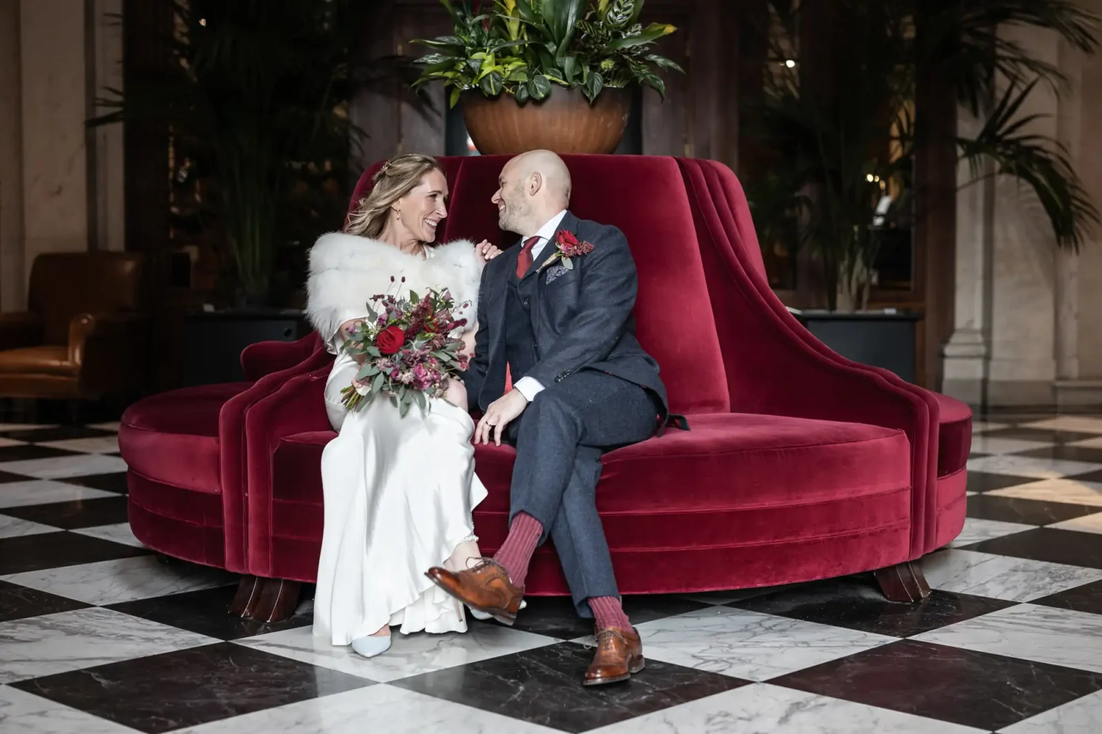 The Edinburgh Grand wedding photographer image of a bride and groom sit on a red velvet couch, smiling at each other. The bride holds a bouquet of red and pink flowers. The setting is a stylish room with a checkered floor.