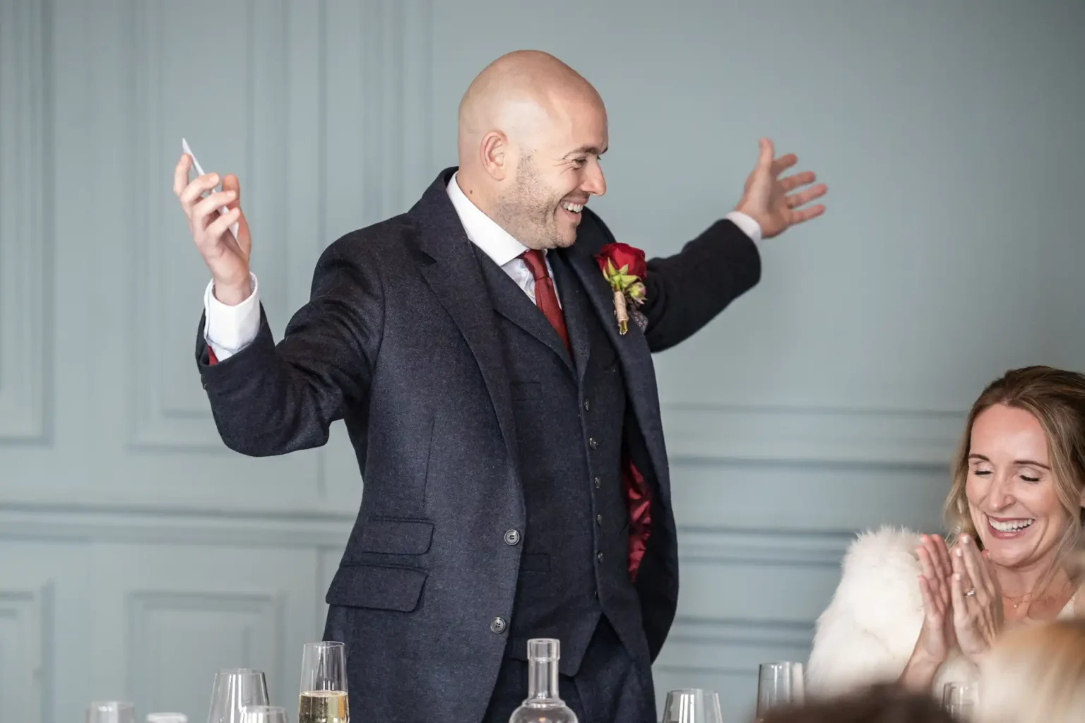 A man in a suit stands with arms wide open, holding a phone. A woman next to him is clapping and smiling.