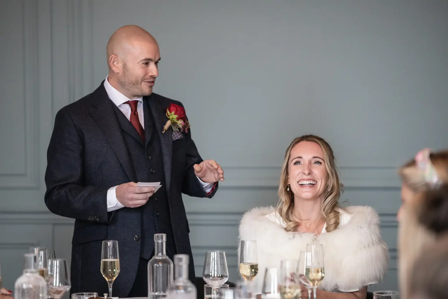 A man in a suit gives a speech next to a smiling woman in a white wrap seated at a table with glasses and bottles.