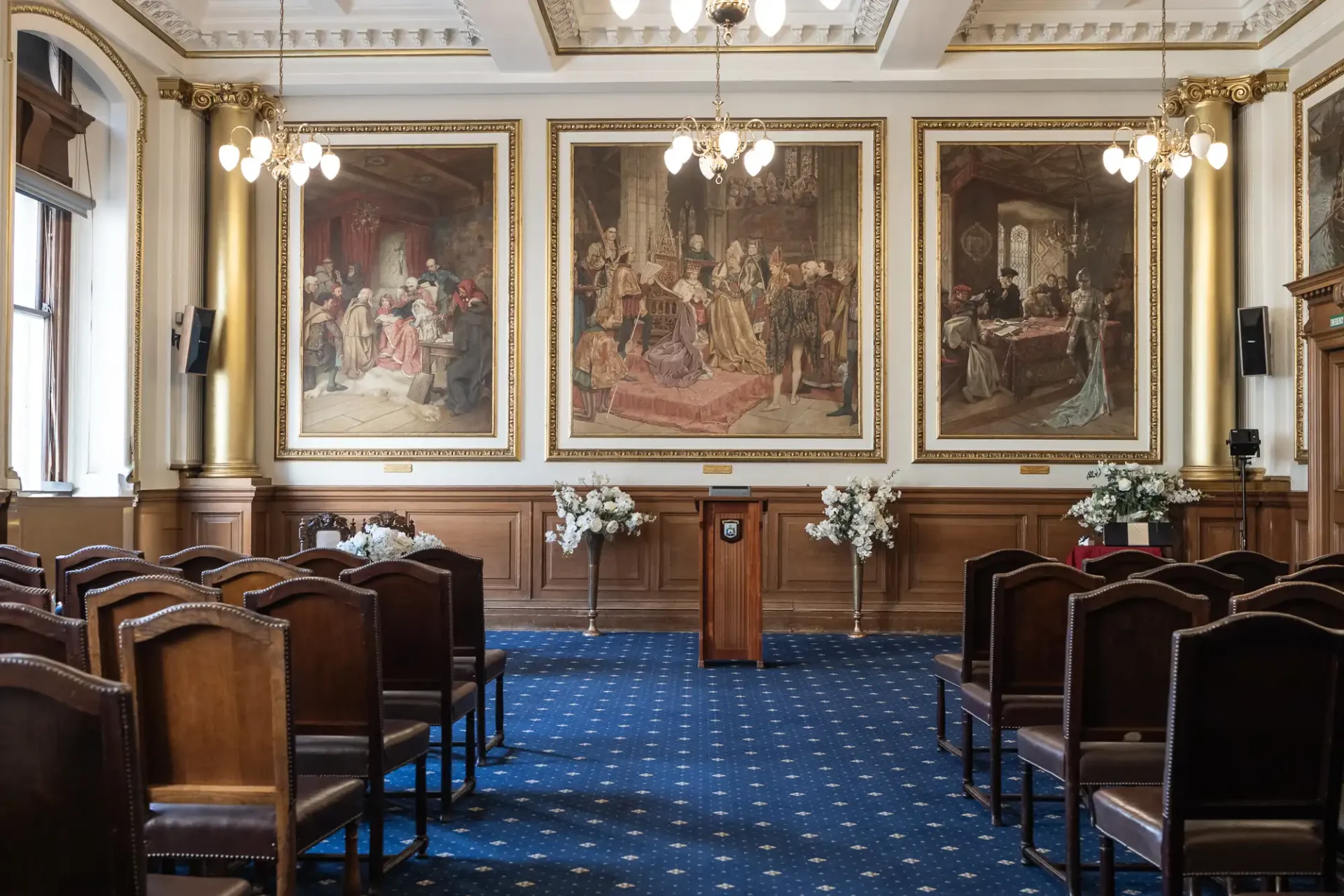 European Room wedding ceremony setup at Edinburgh City Chambers. A room with wooden chairs, a podium, and three large paintings on the wall. Chandeliers hang from the ceiling, and floral arrangements are placed beside the podium.