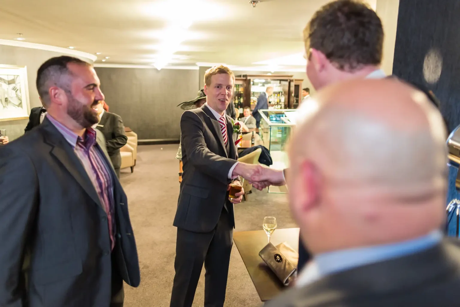 Two men shaking hands at a business event, surrounded by other attendees in a warmly lit room.