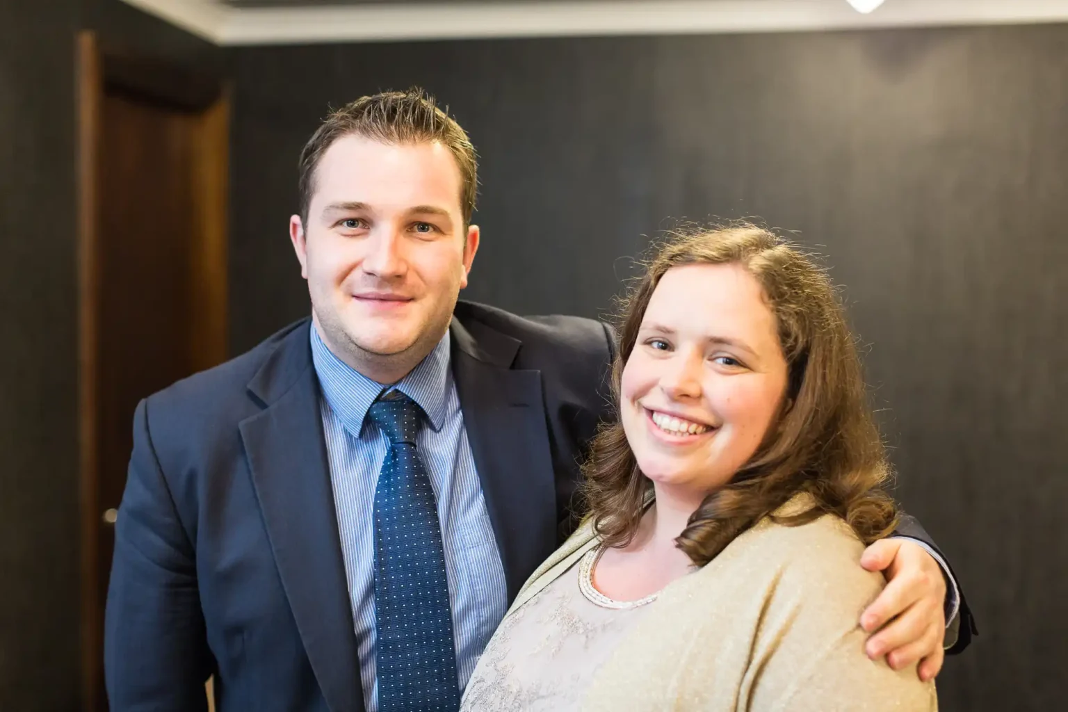 A man in a suit and a woman in a beige cardigan smiling together in a room with dark walls.