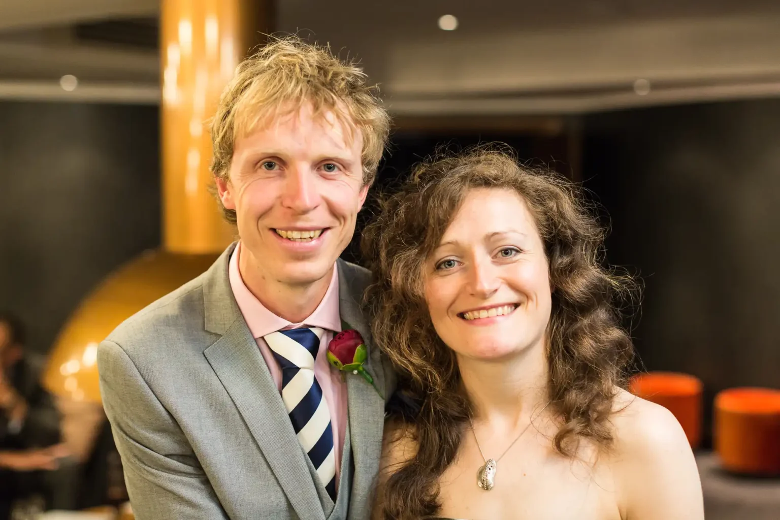 A smiling couple dressed in formal attire posing together at an indoor event, with a man wearing a gray suit and striped tie, and a woman in a white dress with a curly hairstyle.