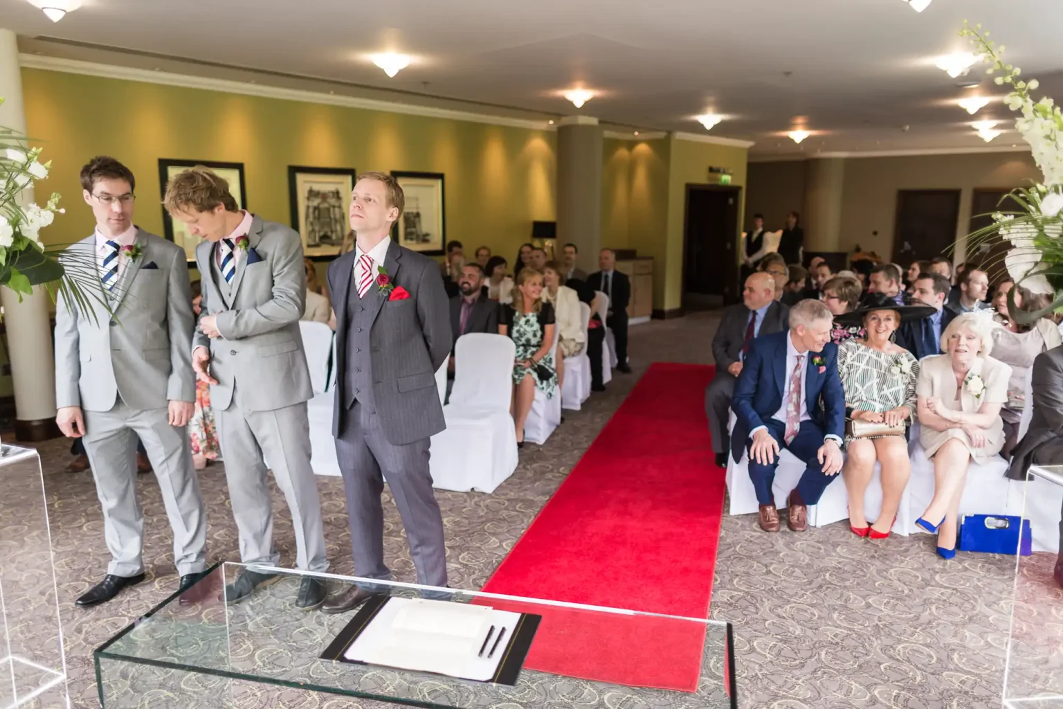 Three men standing at the front of a wedding ceremony room filled with seated guests, one glancing upward with anticipation.