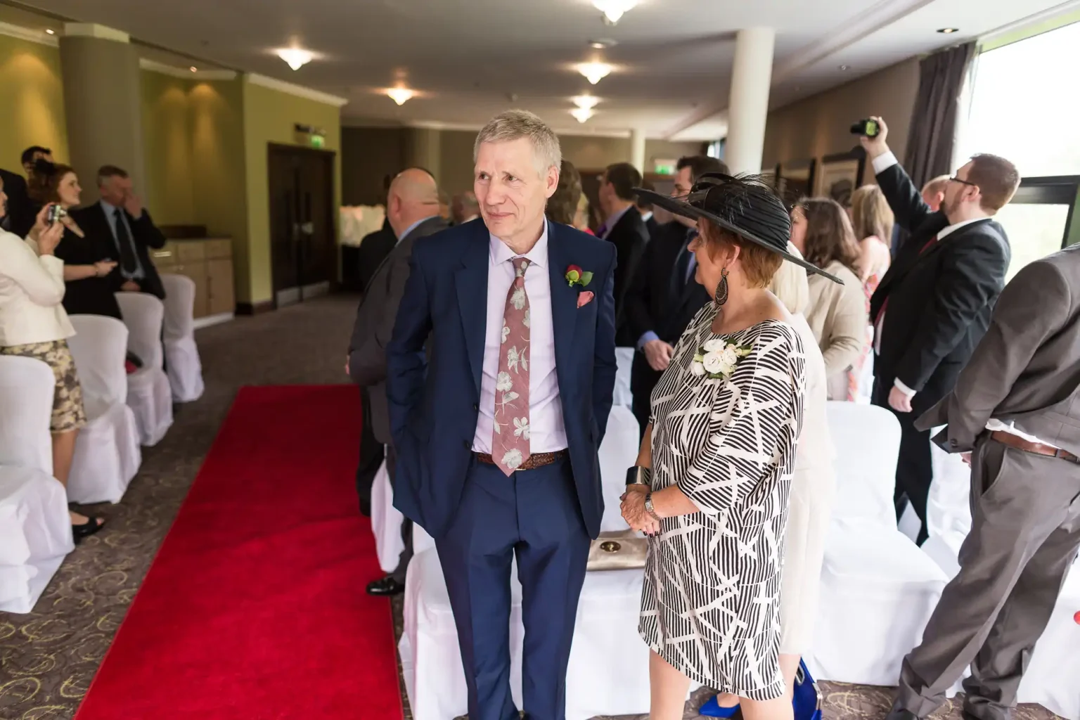 A couple walking down the aisle at a wedding ceremony, guests standing along the sides, with a red carpet pathway.