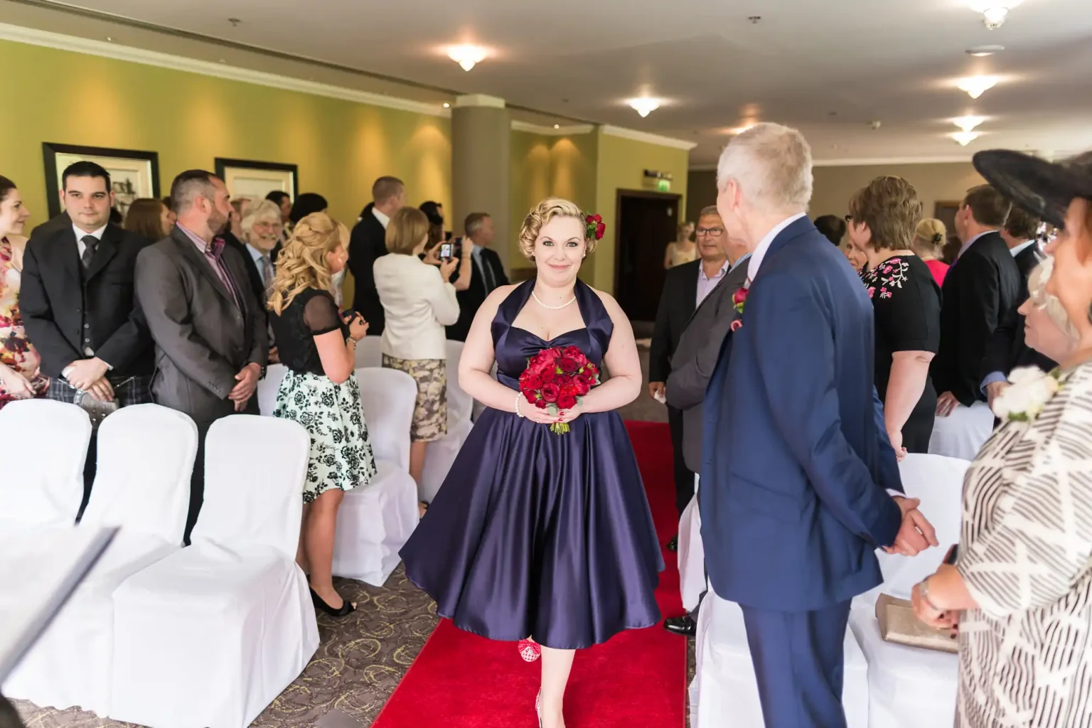 A bride in a purple dress holding a red bouquet walks down the aisle, smiling at the groom who stands at the end, surrounded by guests in a decorated room.