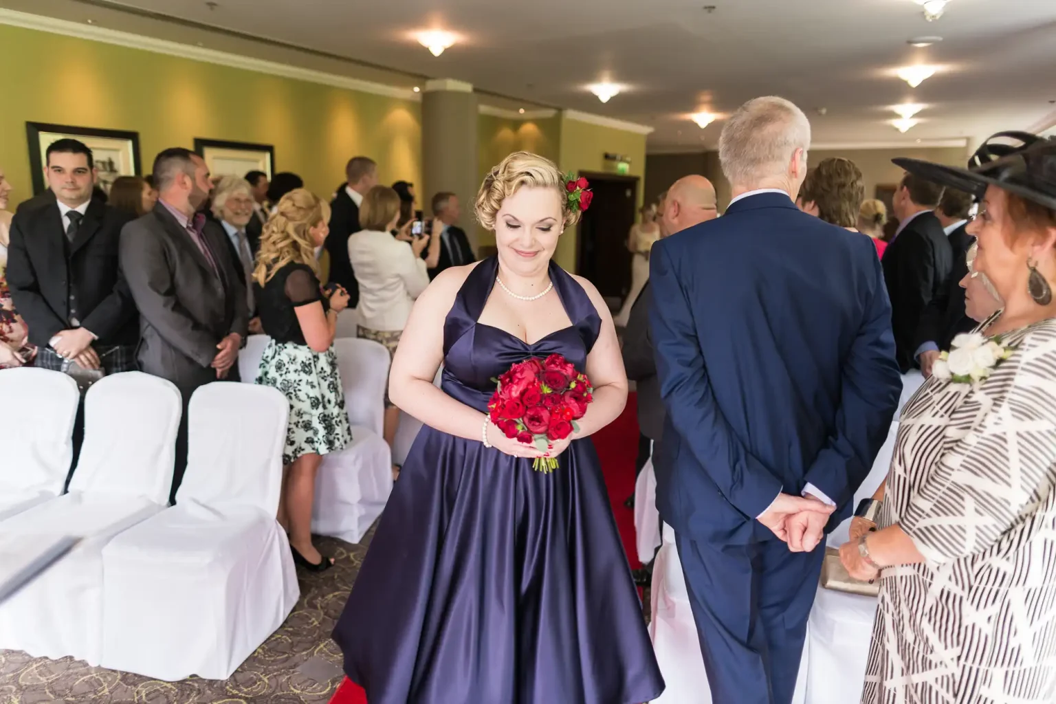 A woman in a purple dress and floral hair accessory holds a red bouquet at a wedding ceremony, surrounded by guests.