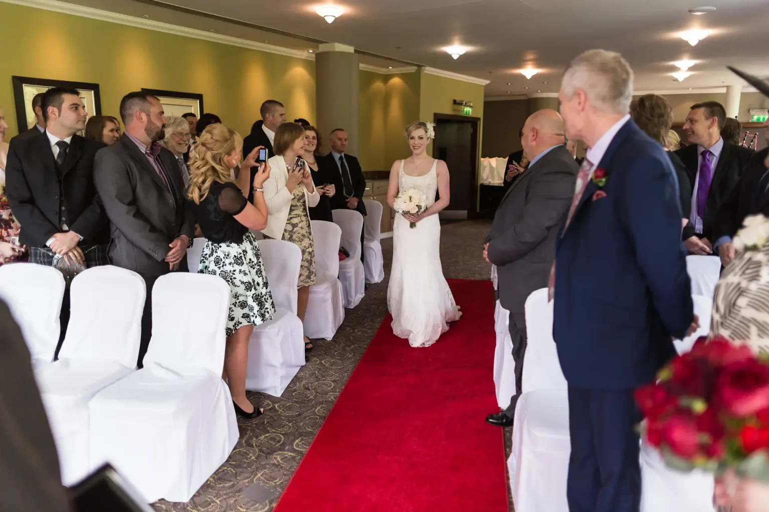 A bride walking down the aisle, holding a bouquet, receives smiles from guests during a wedding ceremony indoors.