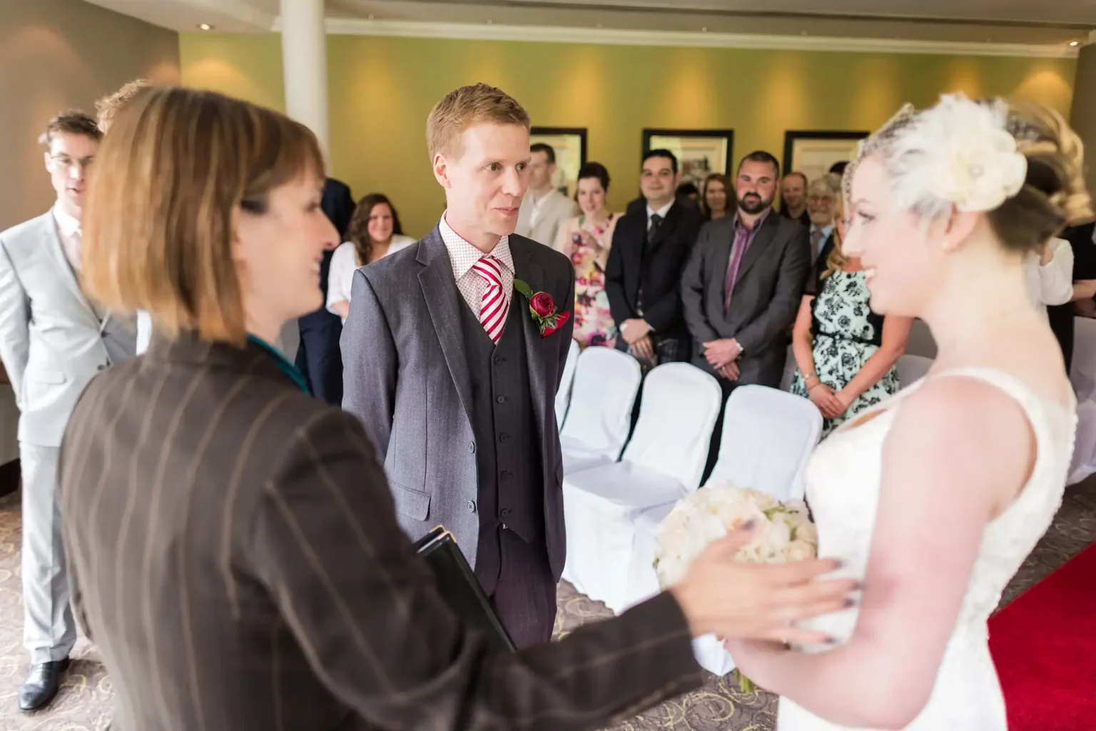 A bride and groom holding hands and facing each other at their wedding ceremony, surrounded by guests in a well-lit indoor setting.