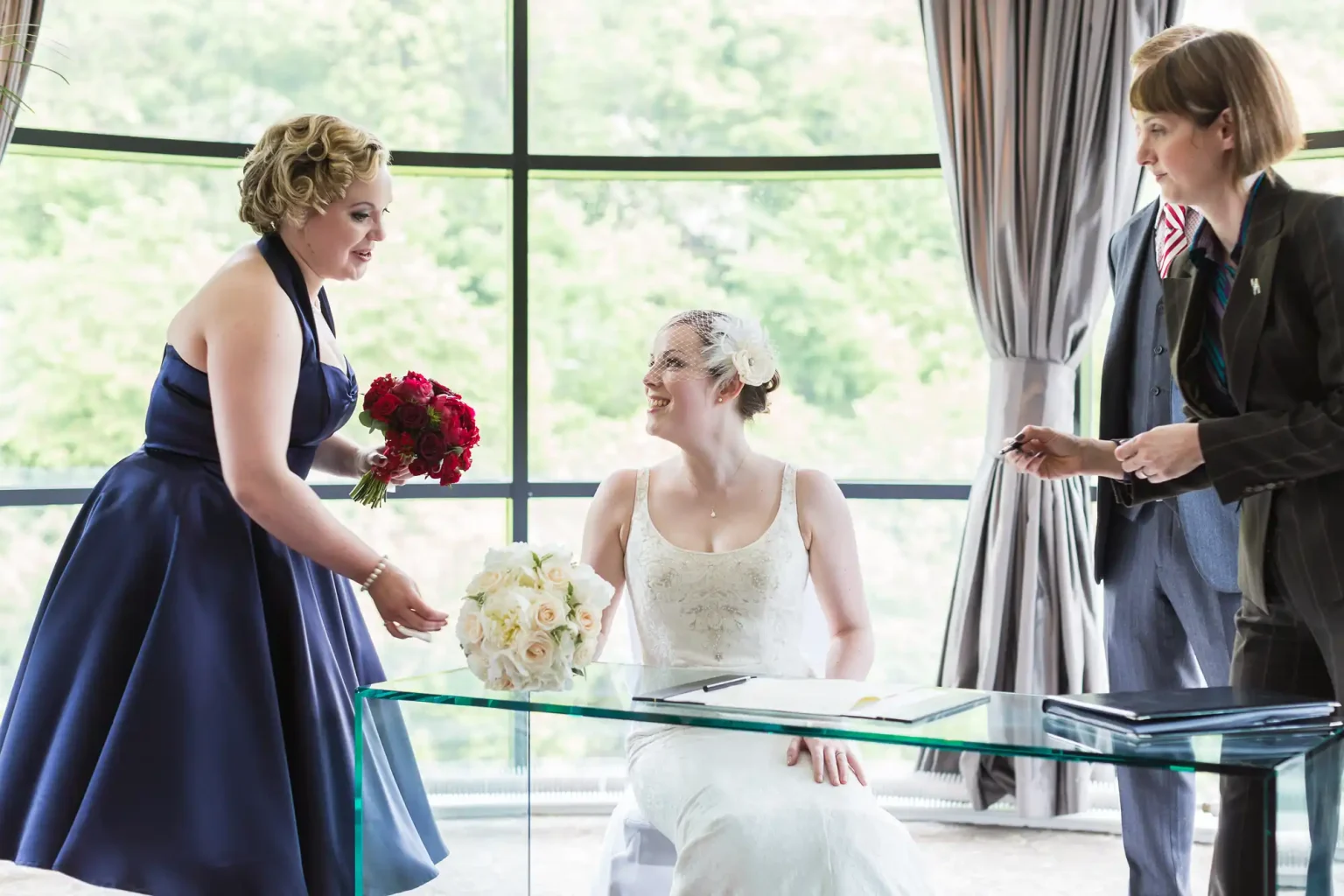 A bride in a white dress and a woman in a blue dress smiling at each other during a wedding ceremony, with an officiant standing by.