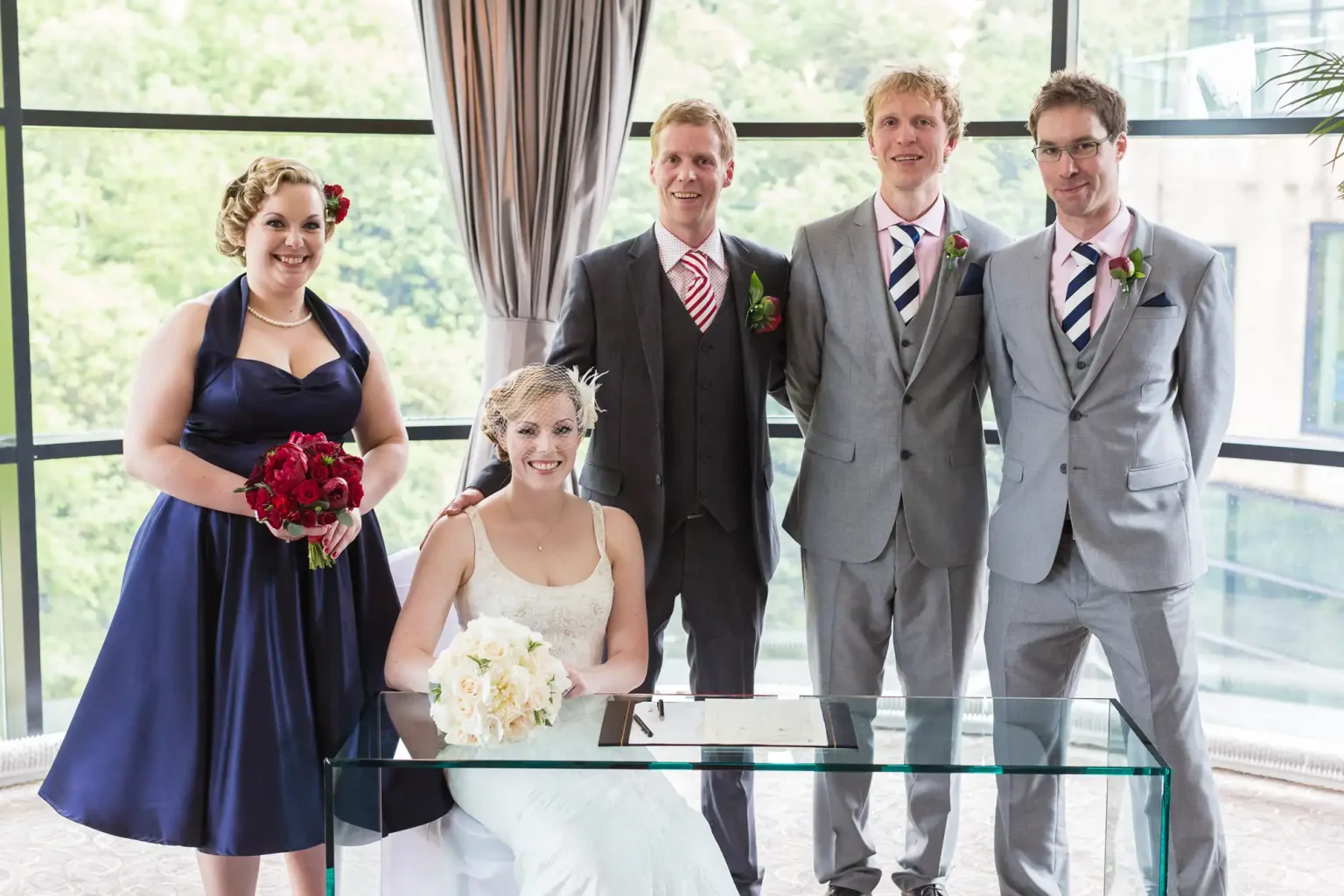 A bride and groom seated at a table with a wedding certificate, flanked by three guests in formal attire, in a room with large windows.