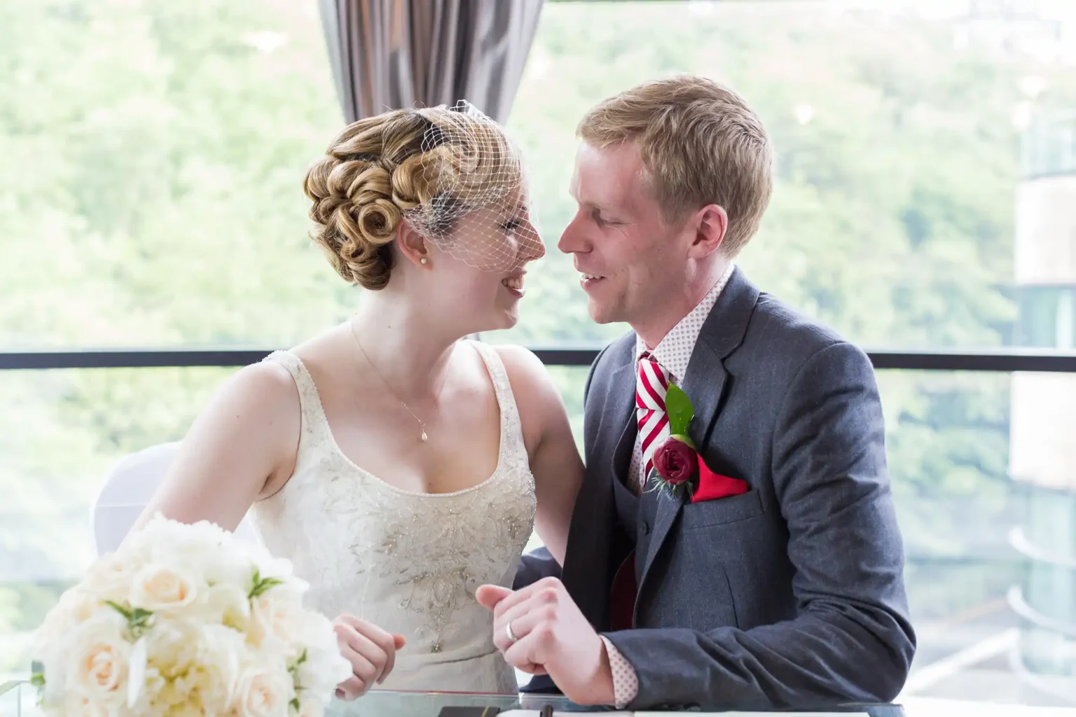 A bride and groom smiling and gazing at each other, with the bride wearing a veil and the groom a red rose boutonniere, indoors by a window.