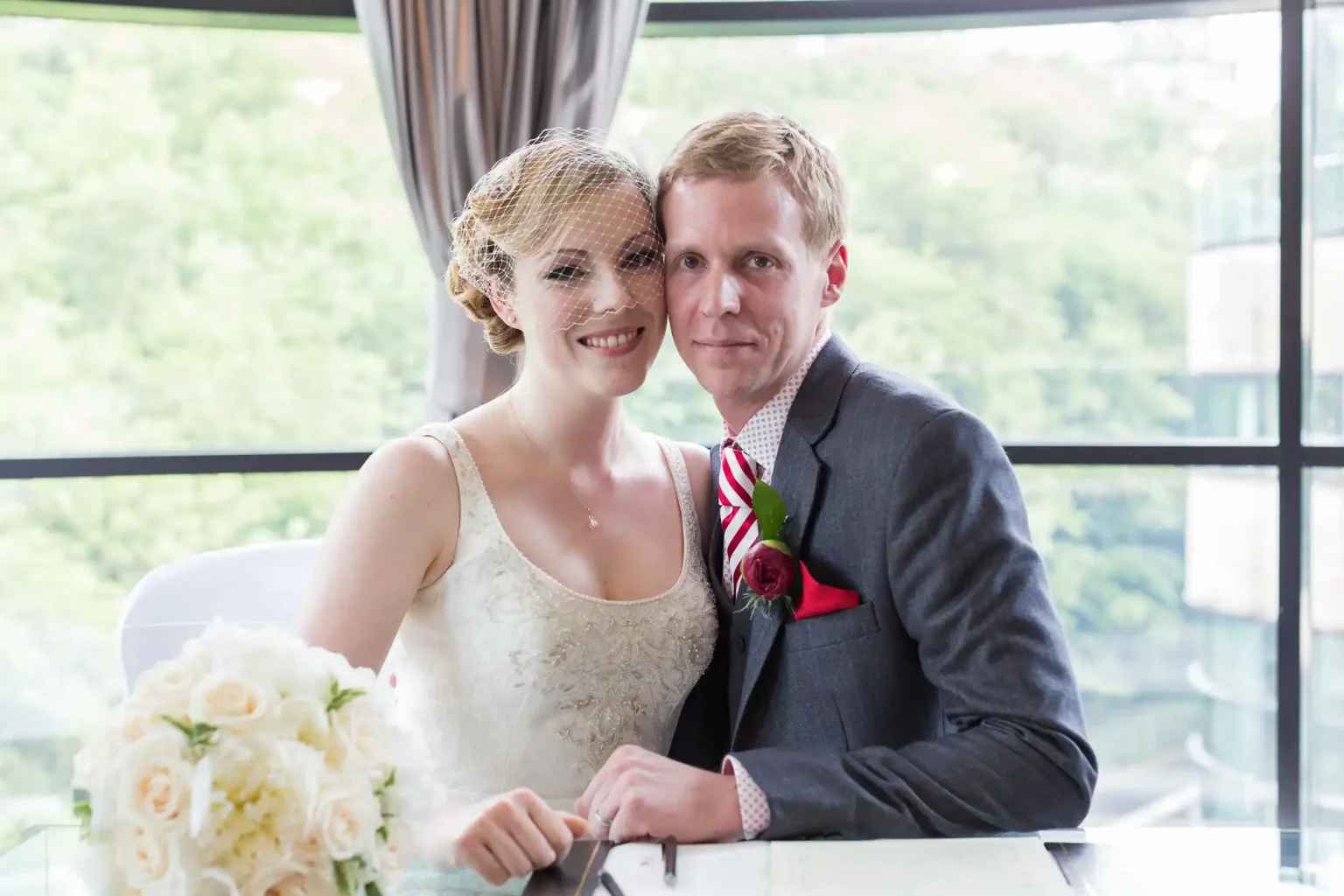 A newlywed couple smiling at the camera while signing their marriage certificate, seated at a table with a floral bouquet in the foreground.