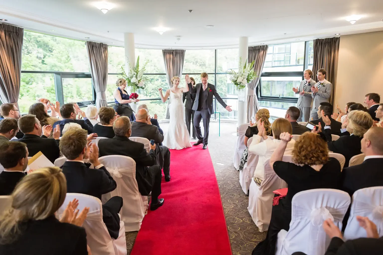 A bride and groom walking down the aisle, applauded by guests, in a well-lit room with large windows and floral decorations.