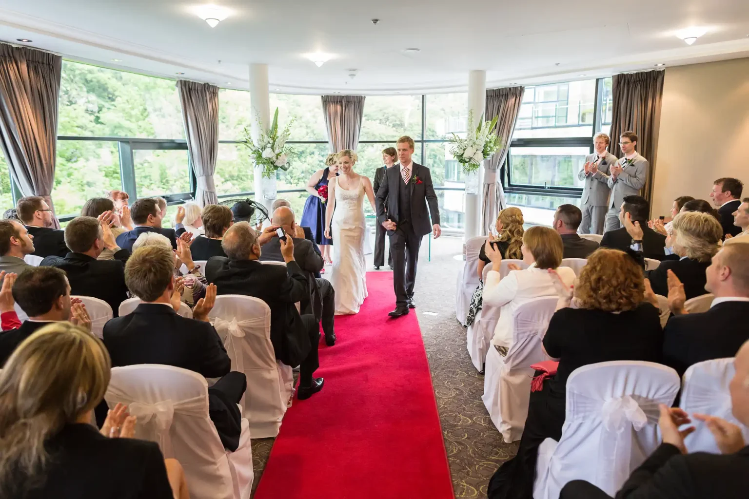 Bride and groom walking down the aisle together, surrounded by seated guests in a well-lit hall with large windows and decorative plants.