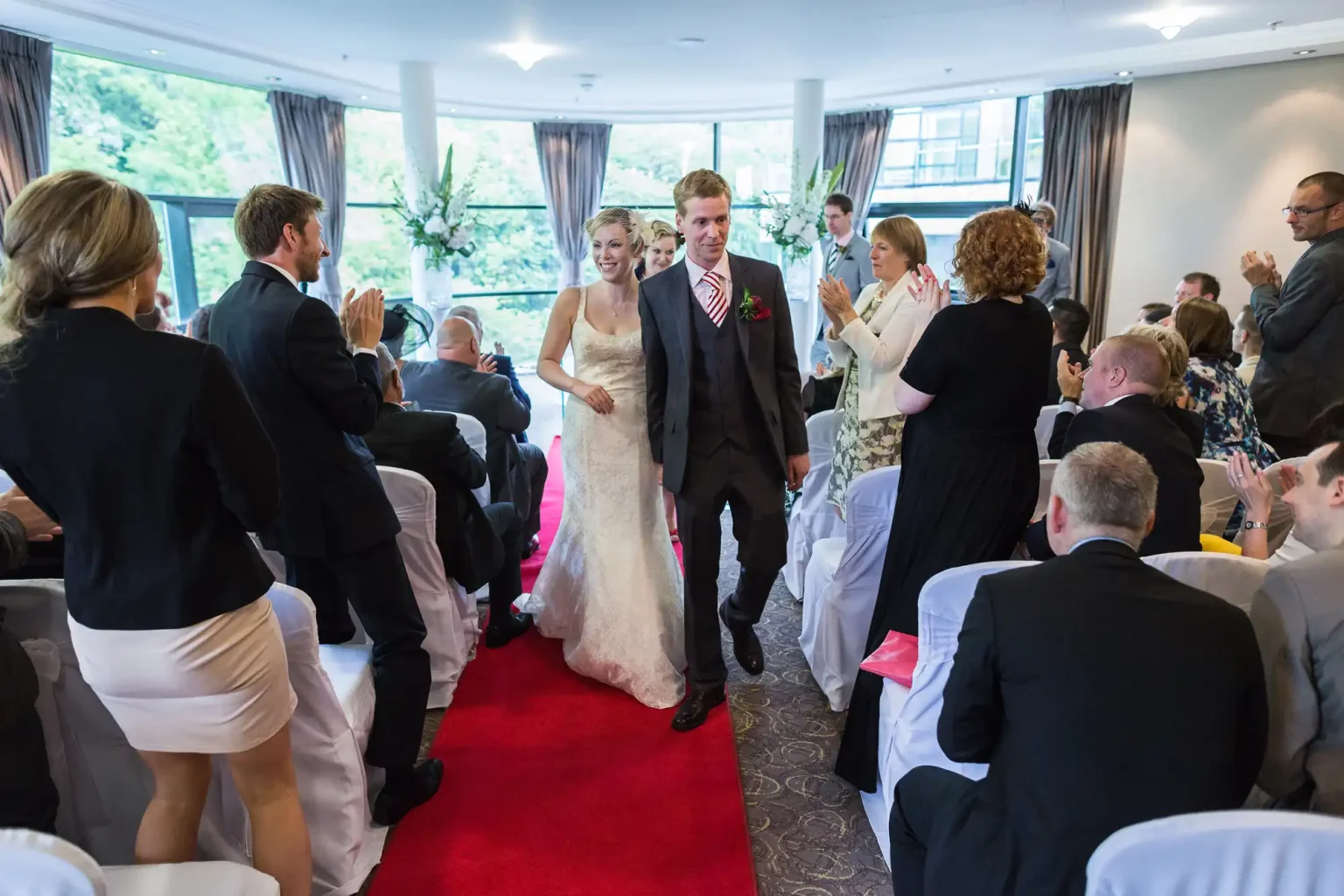 A bride and groom walk down a red carpet, smiling as they exit their wedding ceremony, surrounded by applauding guests.