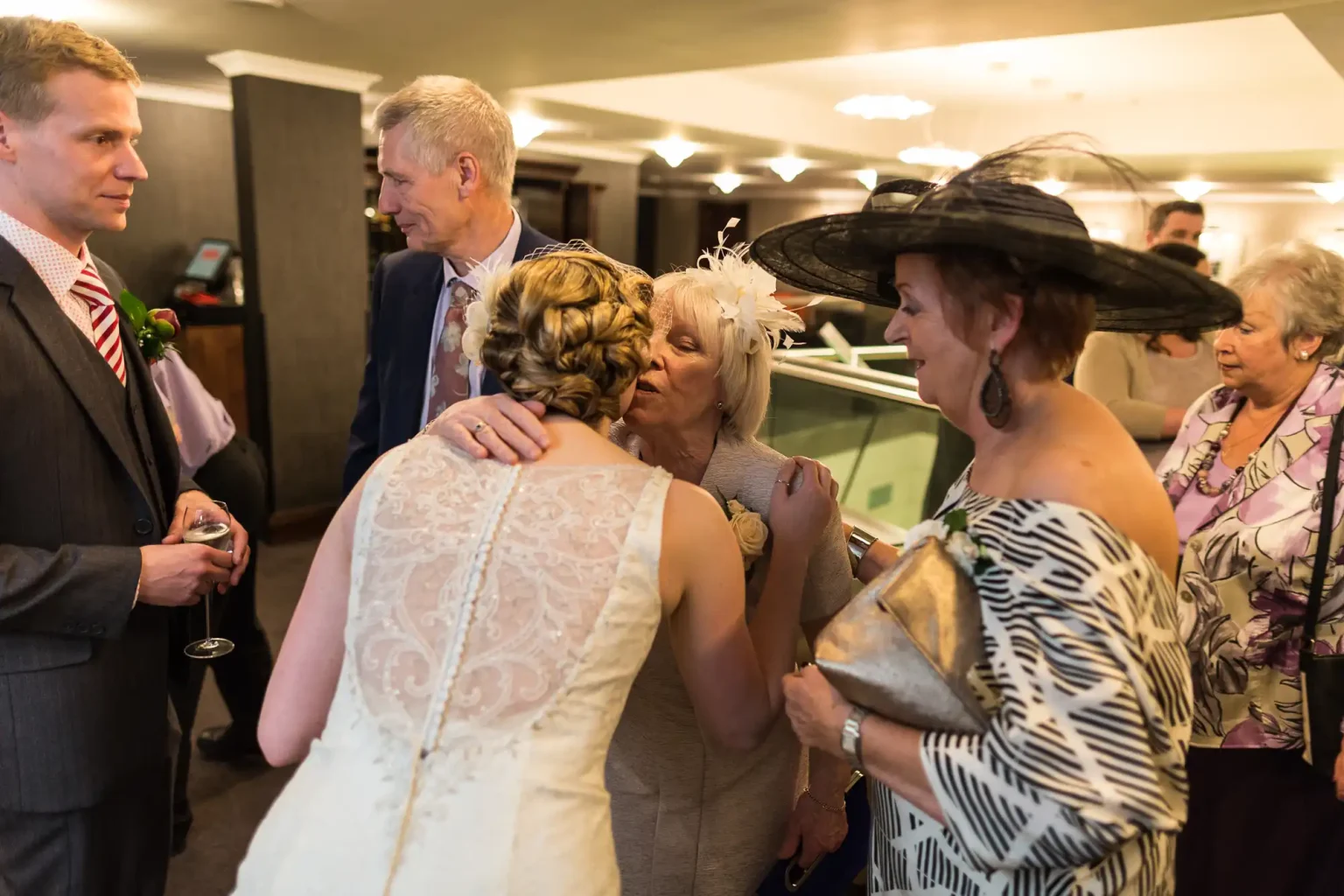 A wedding scene with guests in formal attire chatting indoors; a bride in a lace gown is seen from the back.
