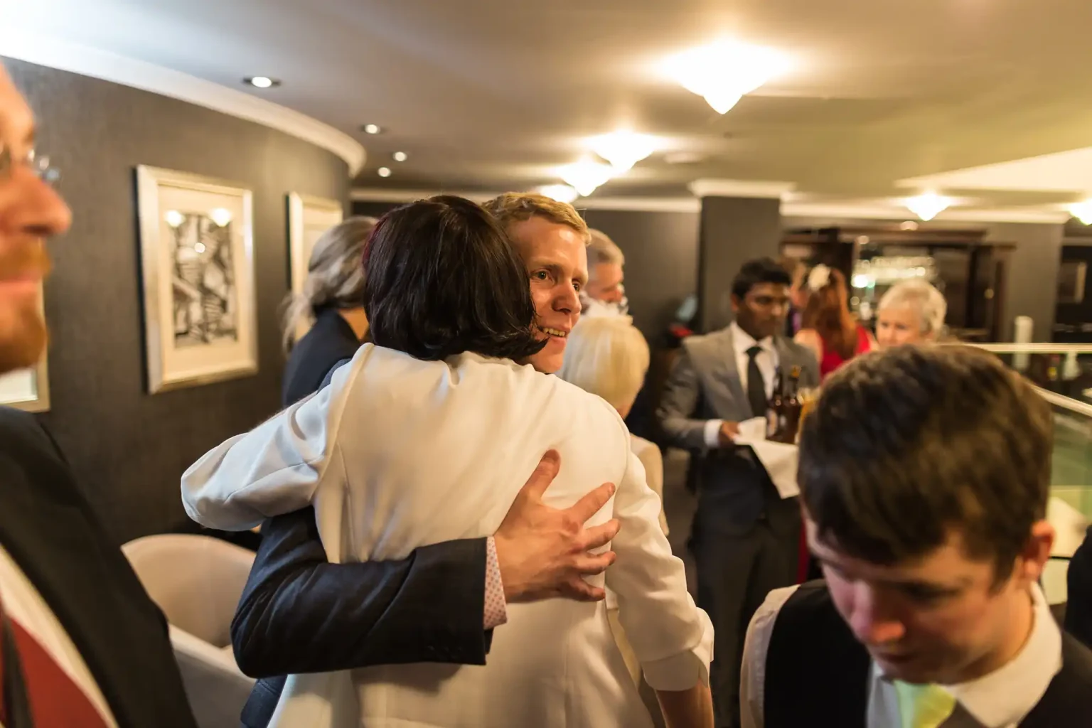 Two individuals warmly embracing at a formal gathering, surrounded by several guests in a well-lit room.