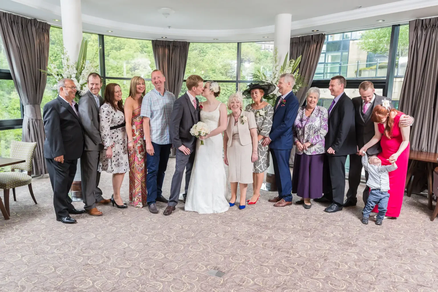 A wedding group indoors, featuring a bride and groom kissing, surrounded by smiling family members of various ages, dressed in formal attire.