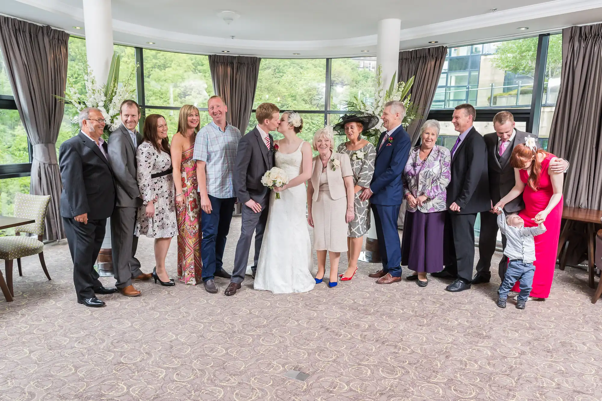 A wedding group indoors, featuring a bride and groom kissing, surrounded by smiling family members of various ages, dressed in formal attire.