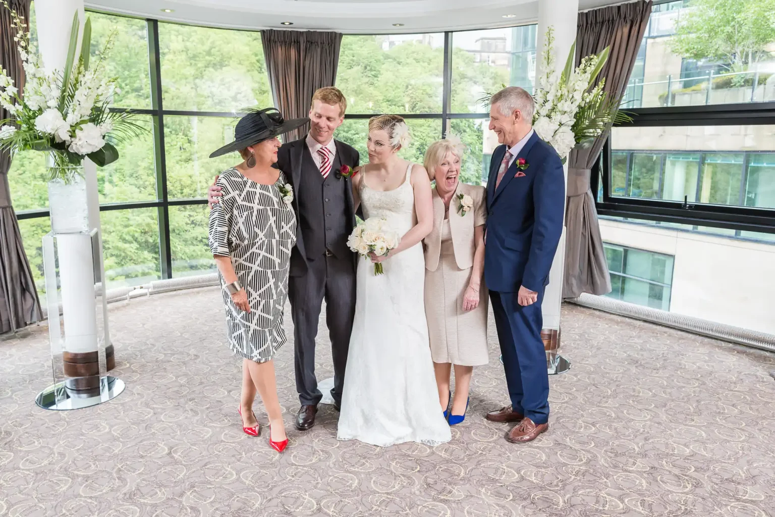 A wedding group standing indoors with five adults--three women and two men--smiling and chatting, surrounded by large windows and floral arrangements.
